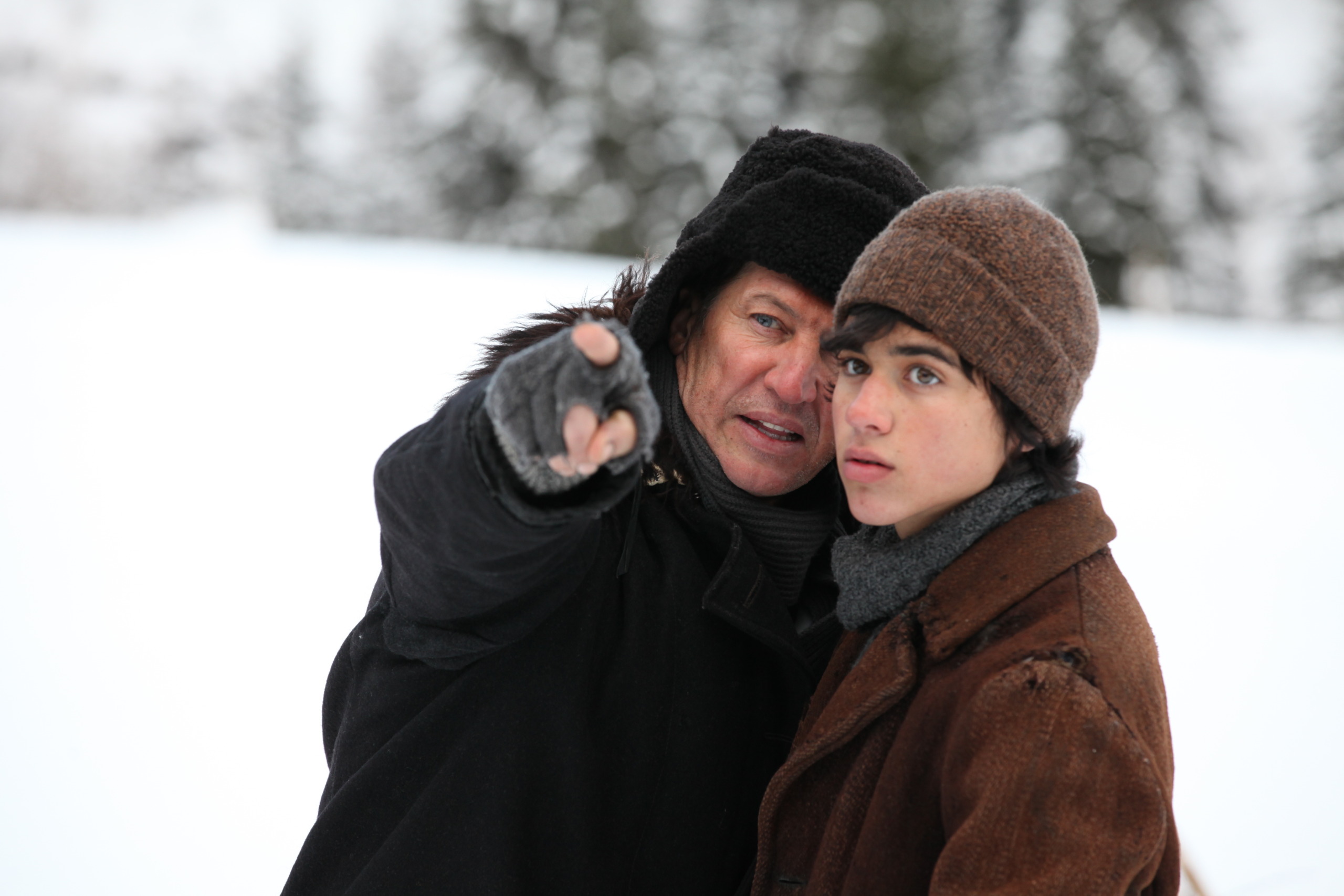 An older man in a warm black coat and hat points ahead, standing next to a younger boy in a brown coat and hat. Together, the pair resemble Brothers of the Wind, venturing through the snowy landscape with blurred trees whispering tales of adventure in the background.