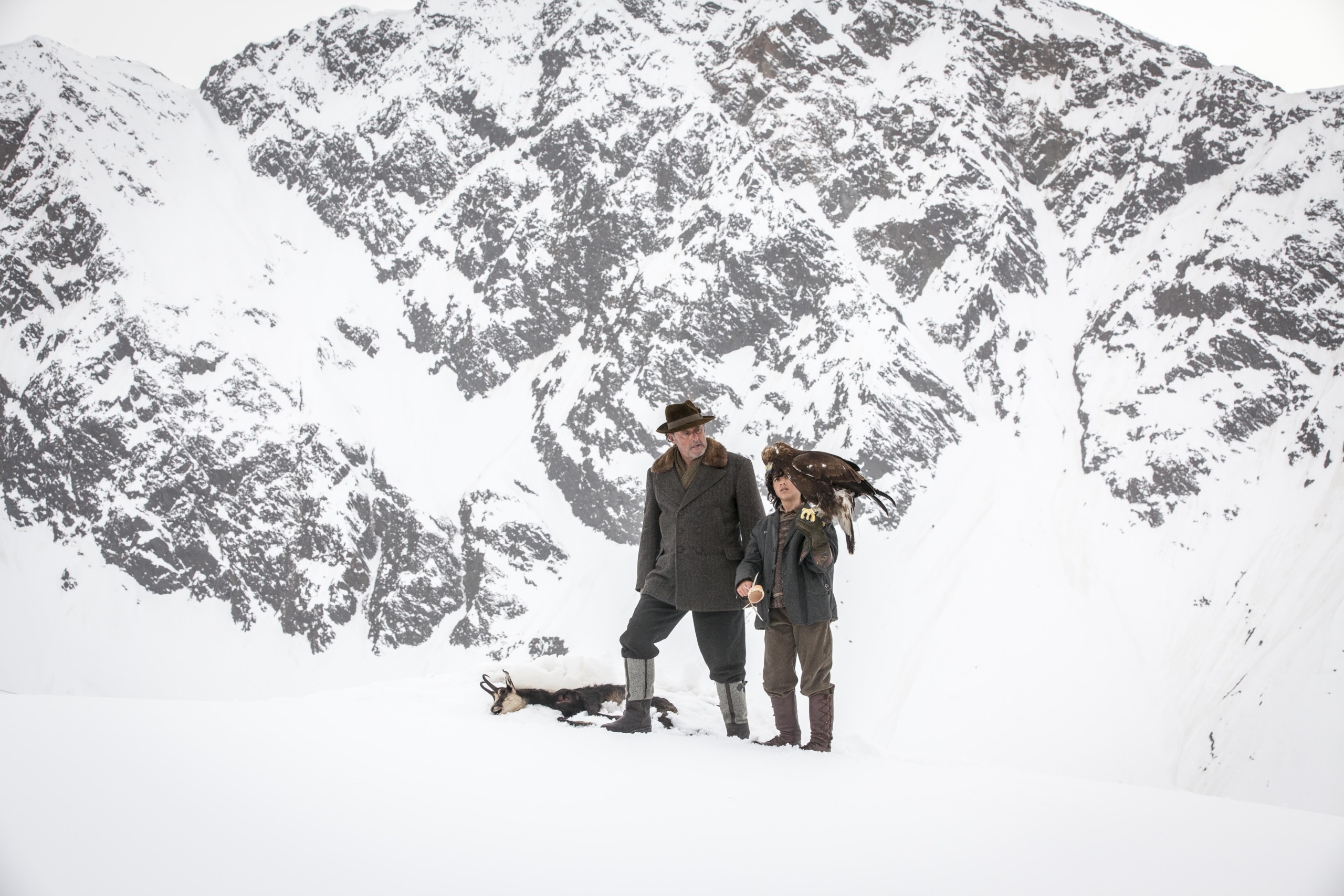 Two brothers of the wind stand on a snowy mountain, with a majestic bird perched on one brother's arm. The other kneels beside a goat resting on the ground, while snow-covered peaks tower in the background.