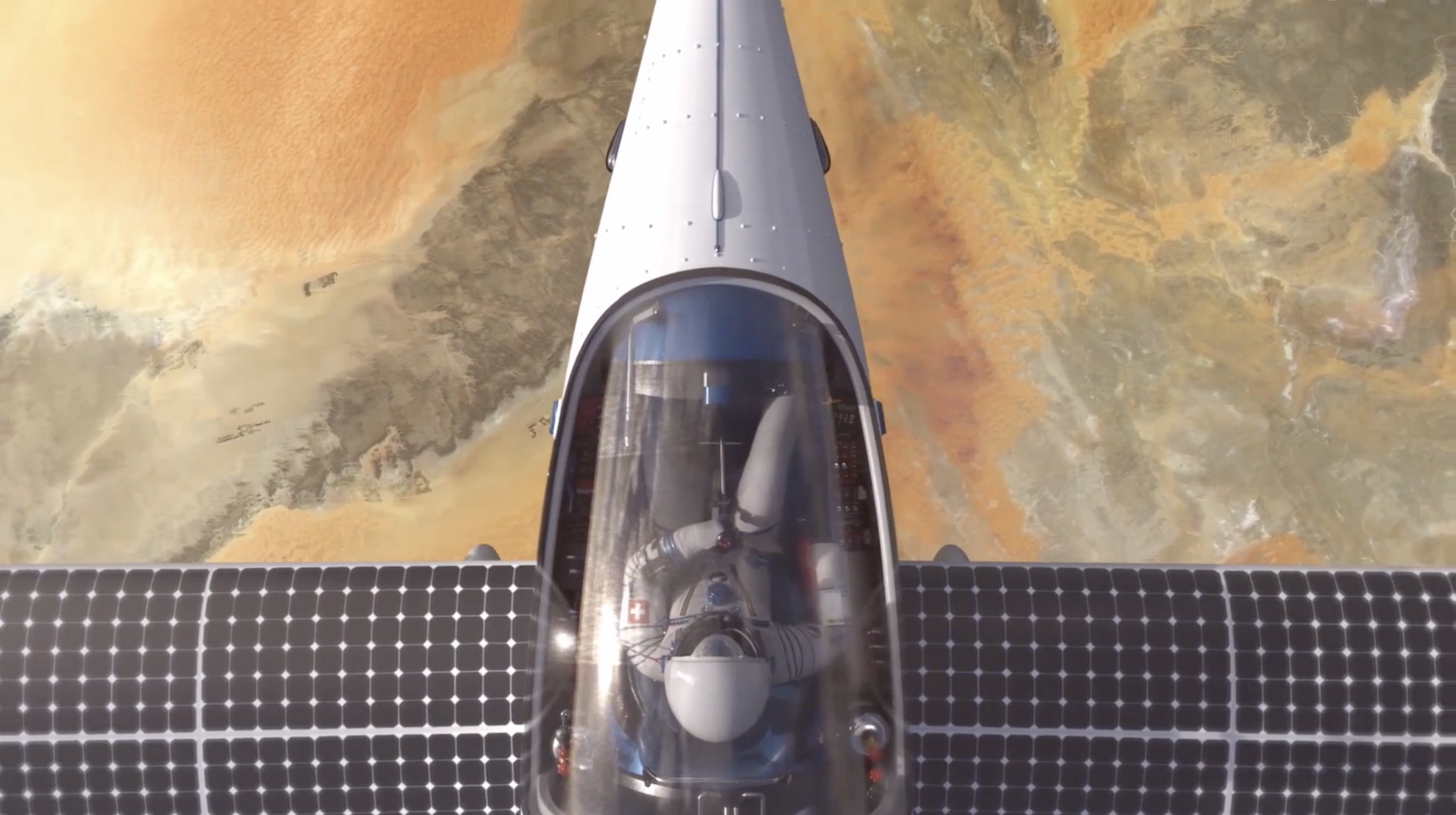 A solar-powered aircraft is flying over a desert landscape. The photo shows a top-down view of the cockpit and the aircrafts wings covered with solar panels. The desert below is a mix of sandy and rocky textures in various shades of brown and orange.