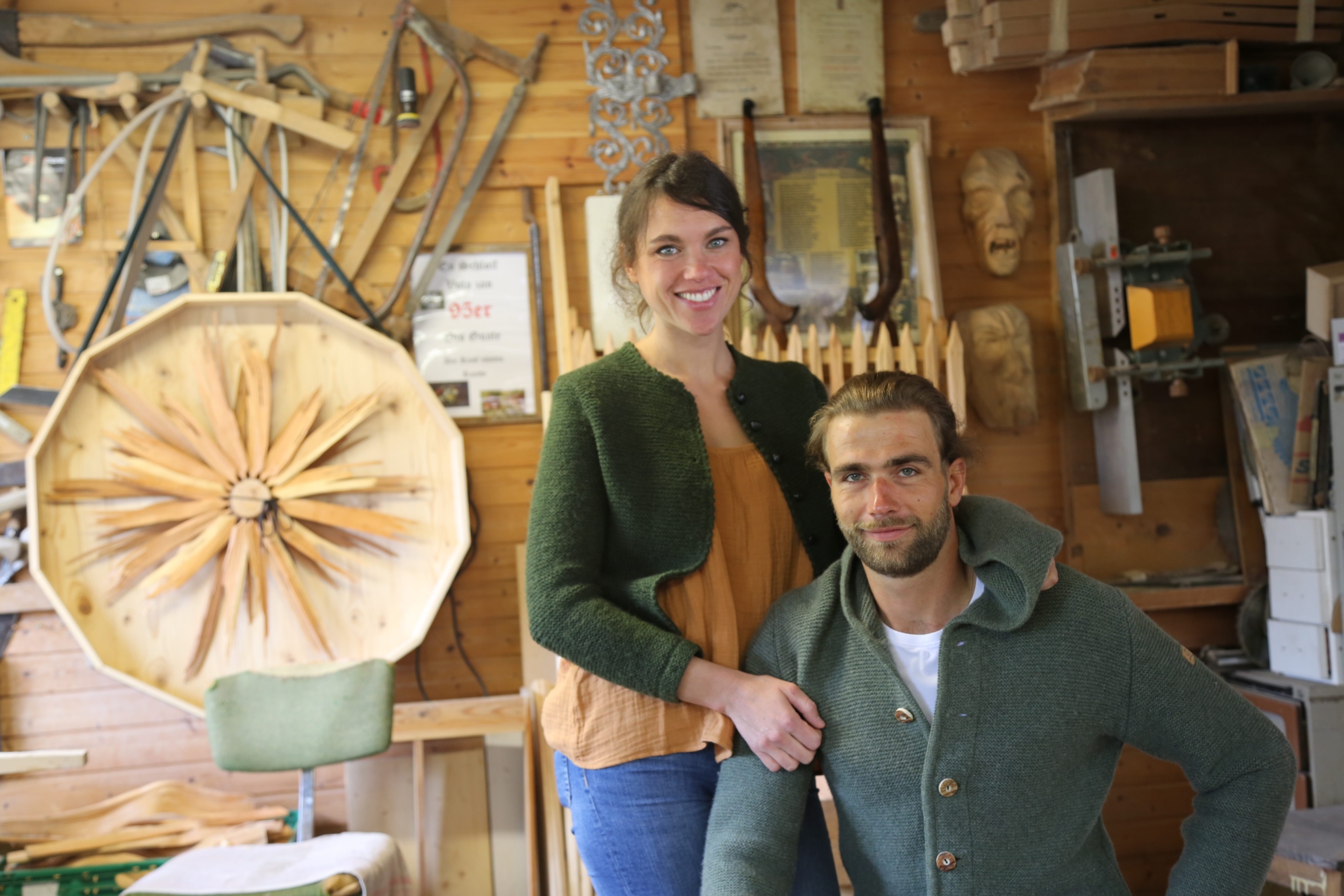 A woman and man smile in a cozy wood workshop, where old traditions meet modern creativity. The space features wooden tools, carvings, and a large decorative piece on the wall. Both wearing green cardigans, they are surrounded by handcrafted wooden items that reflect their wild young spirit.