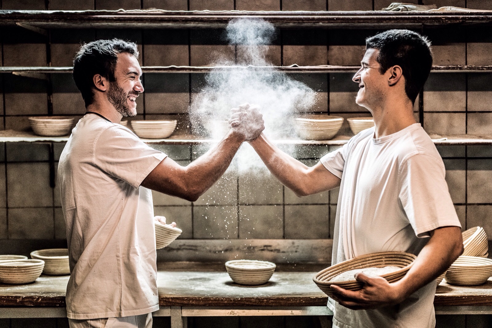 Two bakers in white shirts, holding baskets, joyfully clap hands creating a cloud of flour between them. Embracing old traditions while exuding the spirit of wild young things, they stand in a bakery with shelves lined with wicker baskets, smiling and engaging in playful camaraderie.