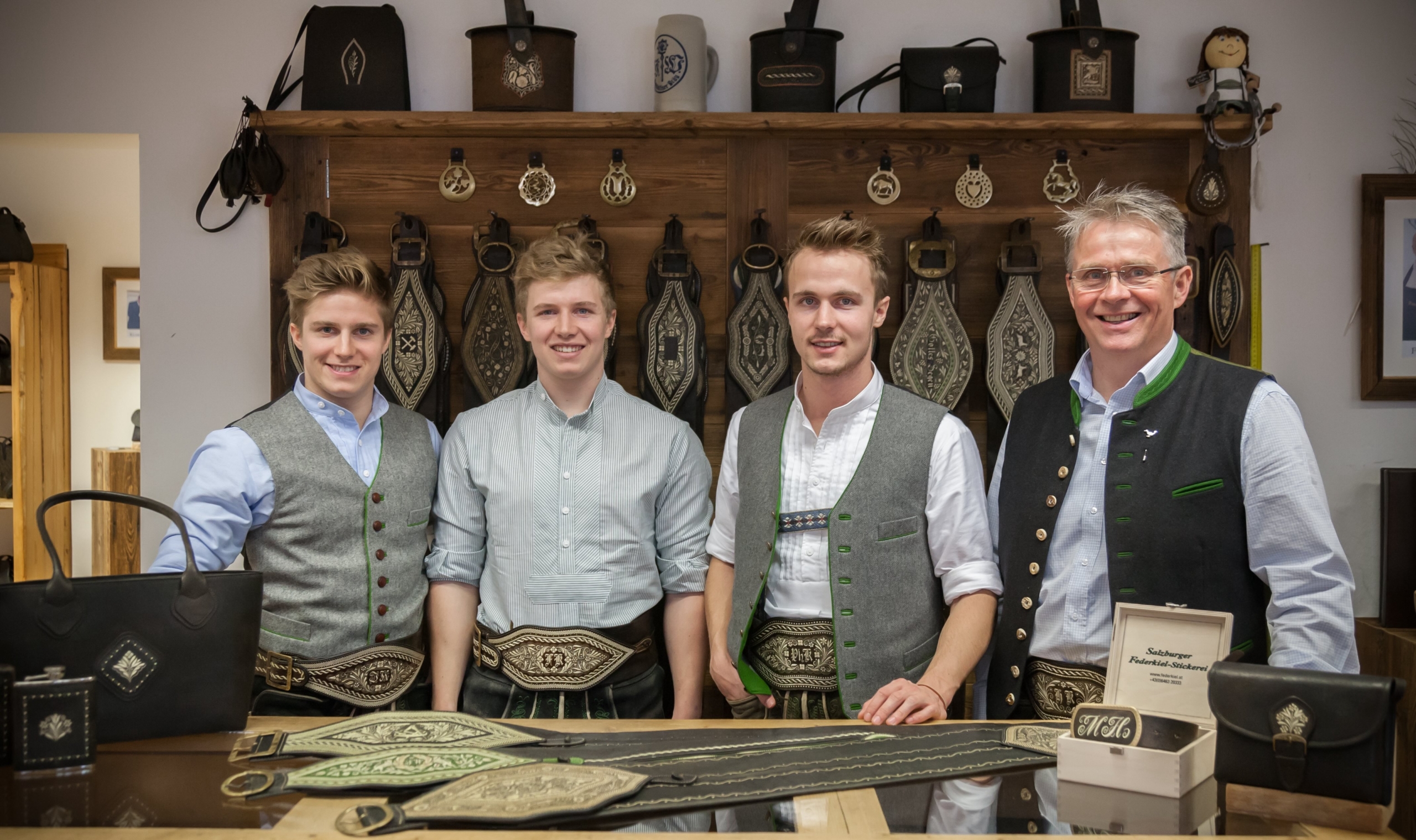 Four men in traditional attire stand in a shop showcasing handcrafted leather goods, embodying old traditions. Behind them, intricate belts and bags are displayed on wooden shelves. They smile warmly, appearing to be part of the store's staff or artisans.