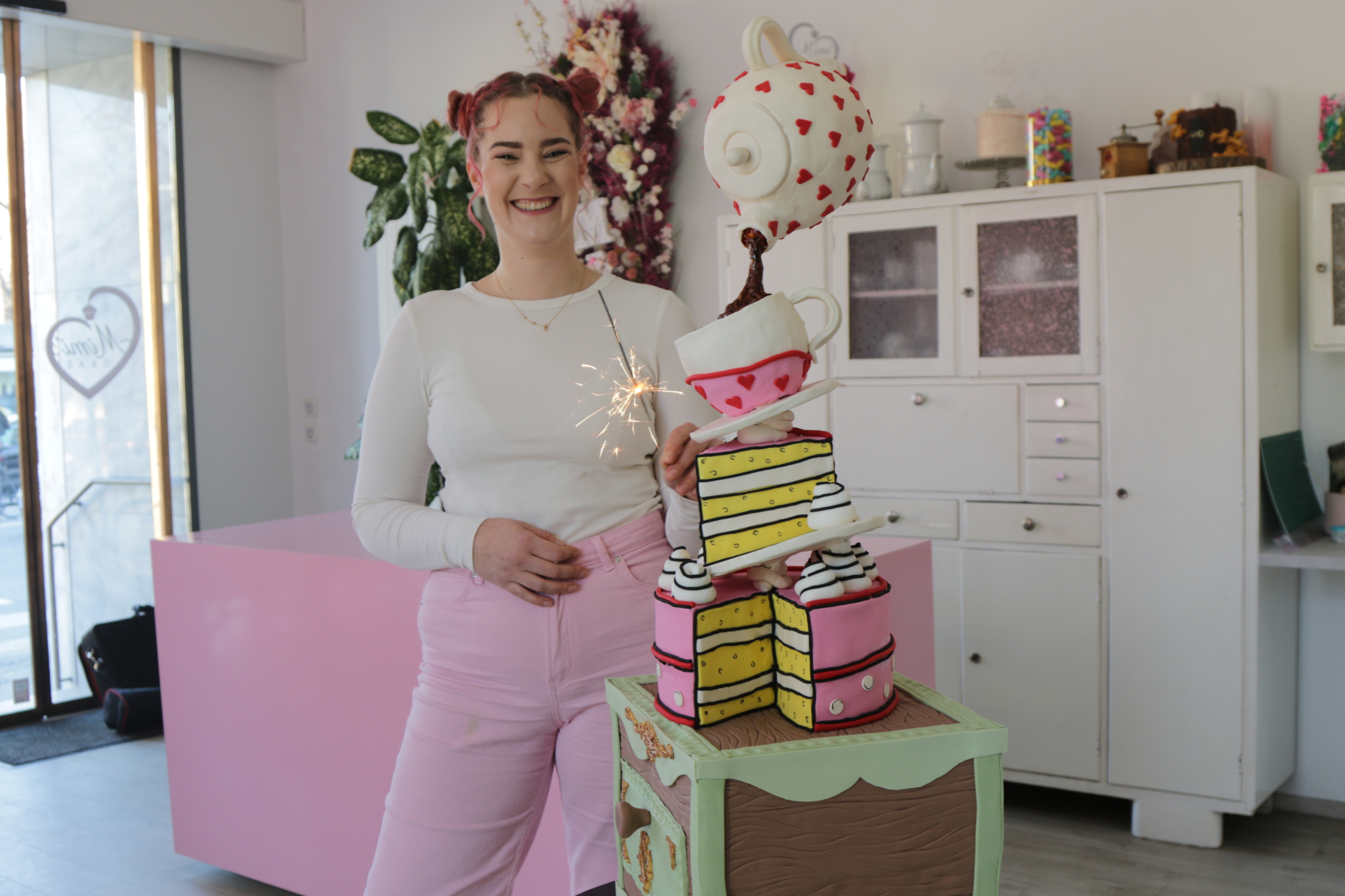 A person with pink hair is smiling while holding a sparkler next to a whimsical cake, crafted by the Wild Young Patissiers. The cake features teacups and striped layers on a decorative stand. The brightly lit room, reminiscent of an Austrian café, is adorned with flowers and cabinets.