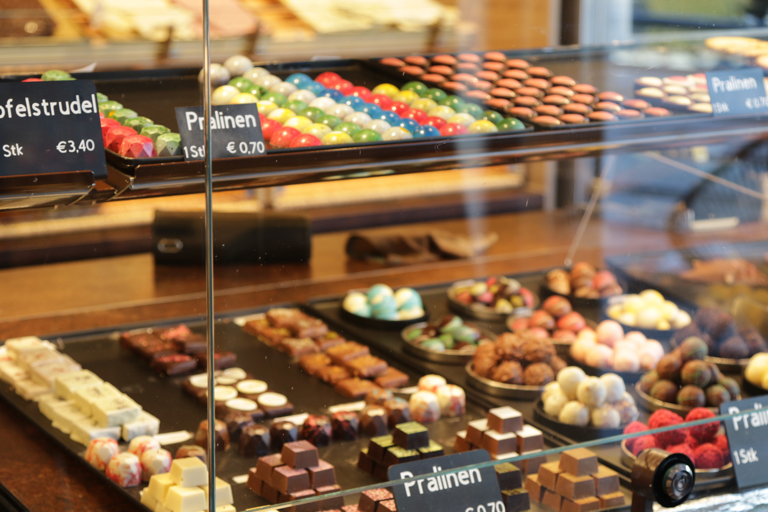 A display case filled with assorted colorful chocolates and sweets crafted by Wild Young Patissiers, including pralines and nougat, each with labeled prices. The treats are arranged in neat rows on dark trays in a well-lit Austrian shop.