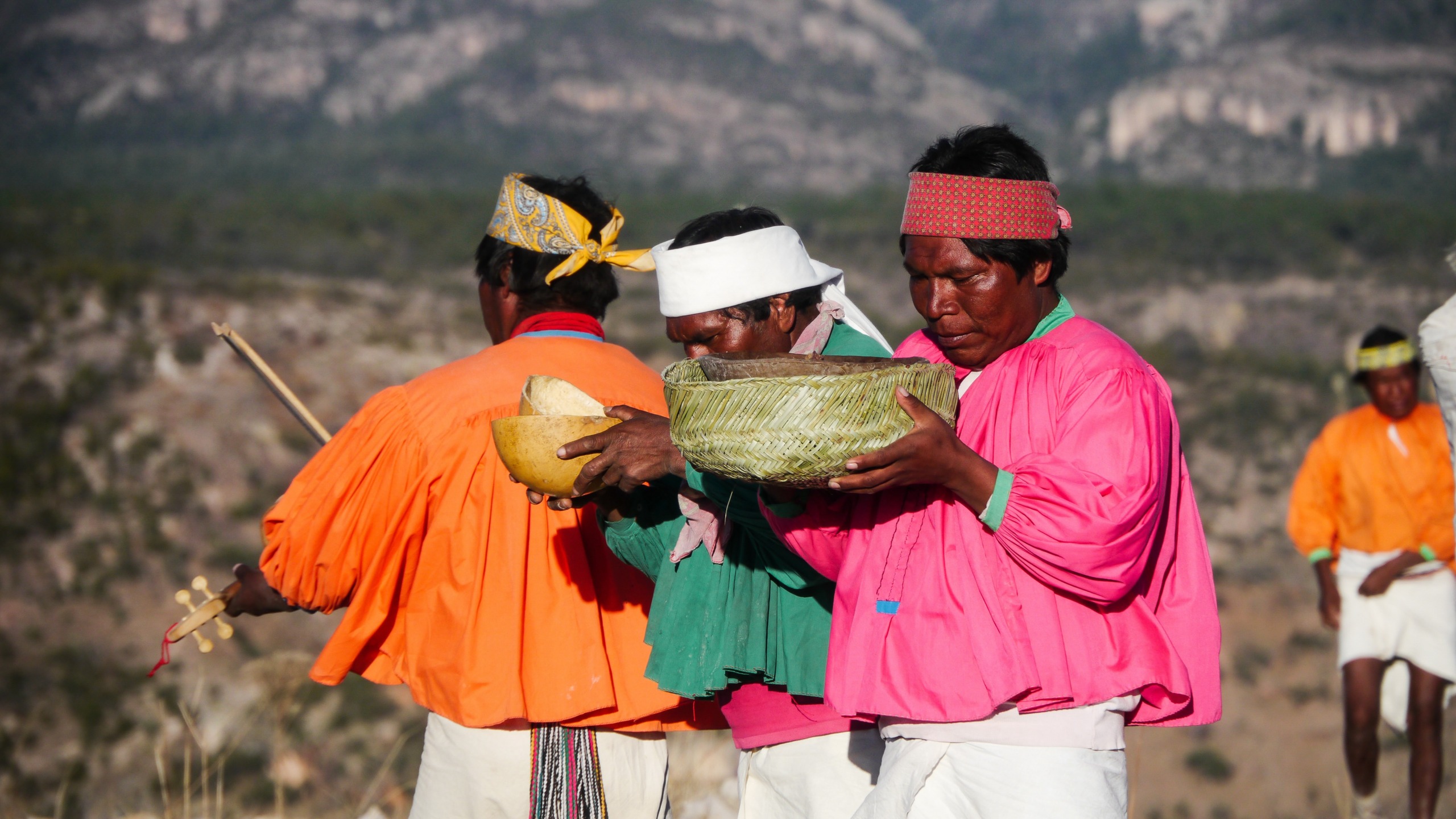 A group of people wearing colorful traditional clothing participate in a ceremony outdoors, embracing the exploration of cultural traditions. They hold large bowls and are focused on the activity, with a mountainous landscape visible in the background.