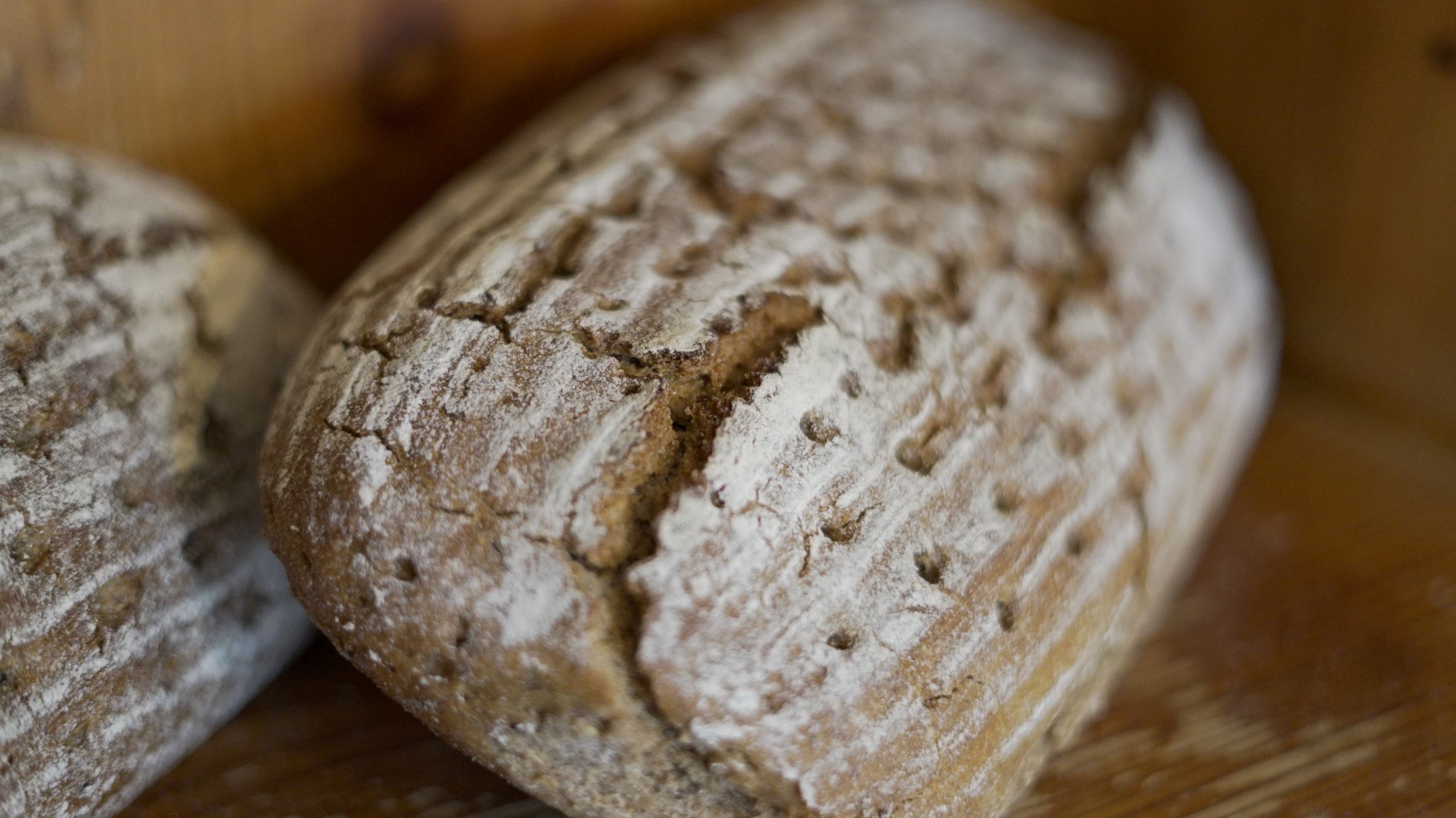 Close-up of a rustic loaf of bread with a cracked, flour-dusted crust, resting on a wooden surface. The texture is highlighted by intricate patterns, truly embodying one's Daily Bread with its artisanal appearance.