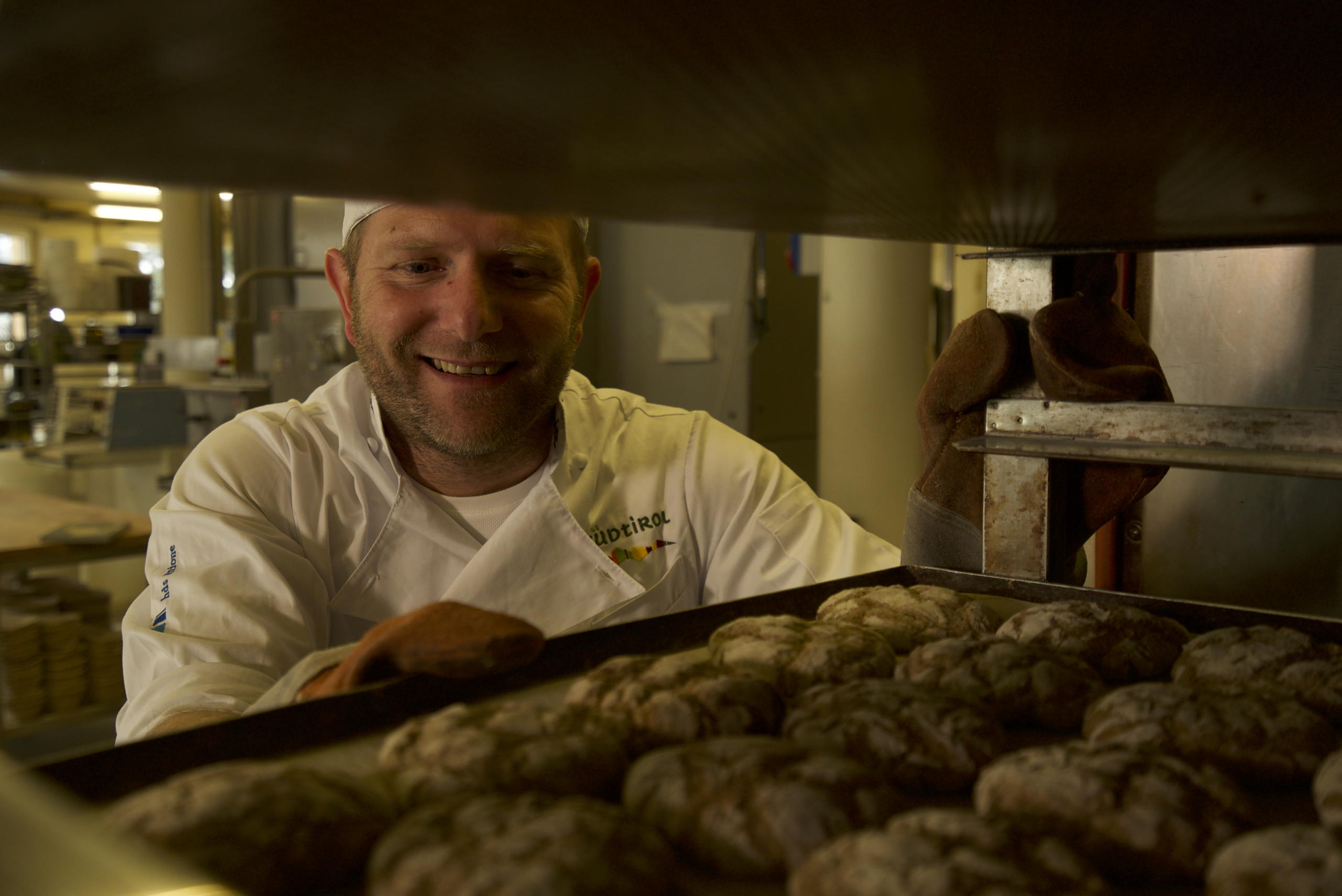 A smiling chef in a white uniform and hat pulls a tray of cookies from the oven, capturing the spirit of Loaf and Soul. Using oven mitts in an industrial kitchen, he embodies the dedication to crafting daily bread with passion and precision.