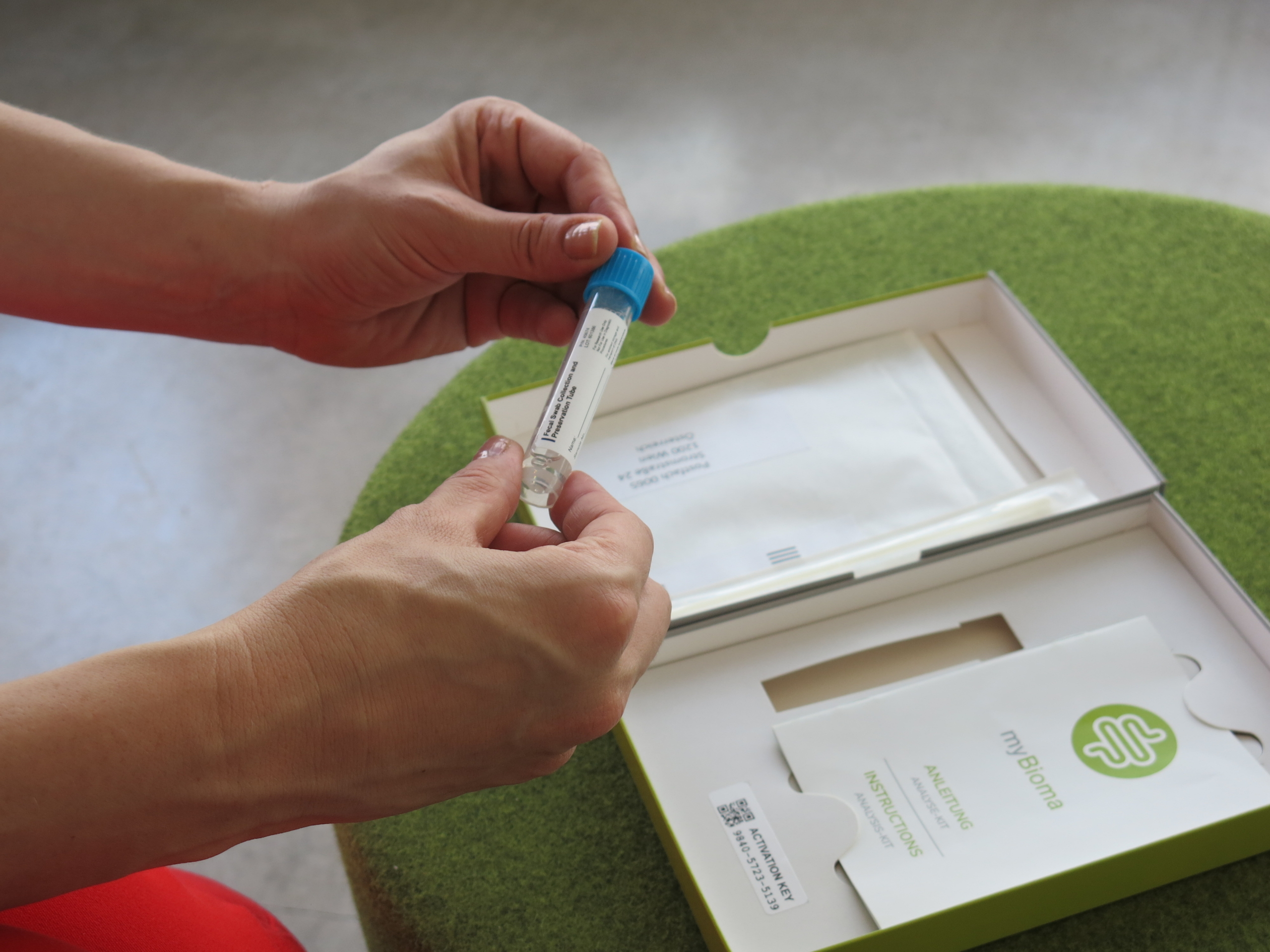 A person holds a plastic test tube with a blue cap, intently observing its contents. This tube is part of a home testing kit for intestinal health, complete with a box and an instruction booklet labeled my biome, all resting on a green circular table amidst myths of gut wellness.