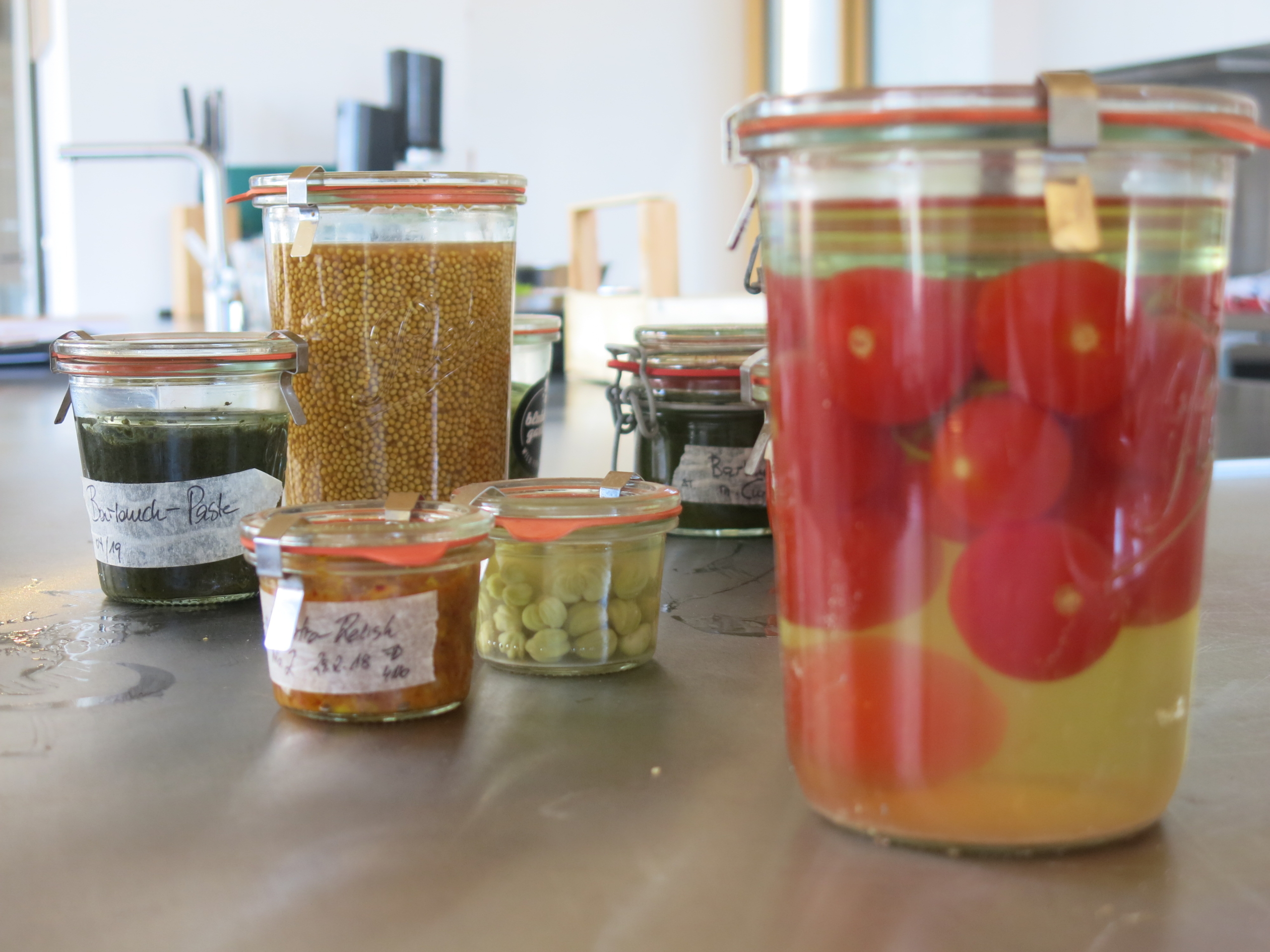 Jars of preserved foods line the kitchen countertop, including tomatoes, intestinal-friendly mustard seeds, peas, and pesto. Each jar is sealed with rubber gasket lids and features handwritten labels that dispel myths about home canning.