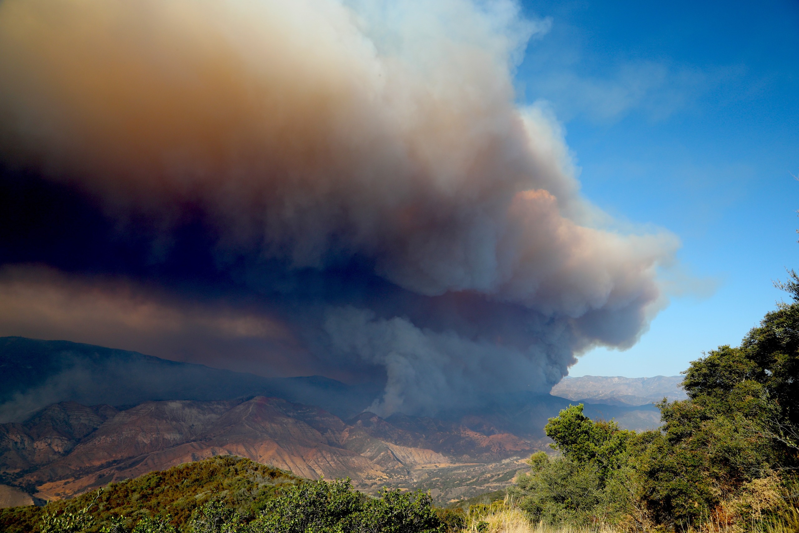 A large, dark smoke cloud billows over the mountainous landscape of California, contrasting with the clear blue sky. Lush green trees are visible in the foreground, highlighting the dramatic scene of a distant wildfire.