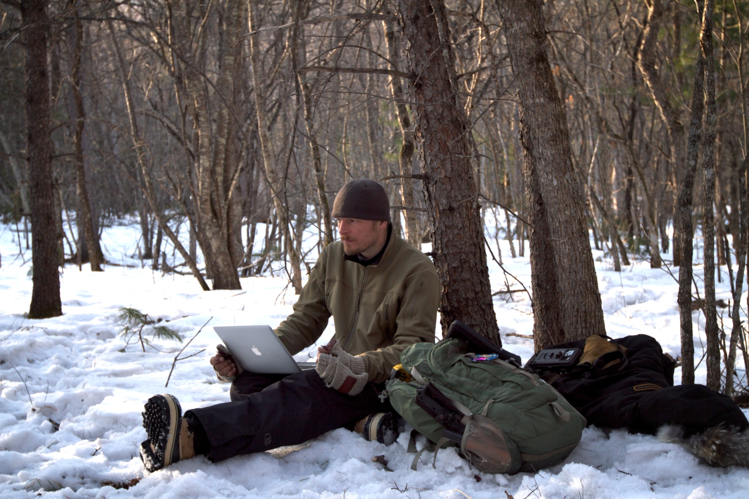 A person sits on snow in a forest, using a laptop. They're wearing winter clothing and a beanie with a tiger pattern. A large green backpack and other gear are nearby. Bare trees surround them, and the ground is covered in snow.