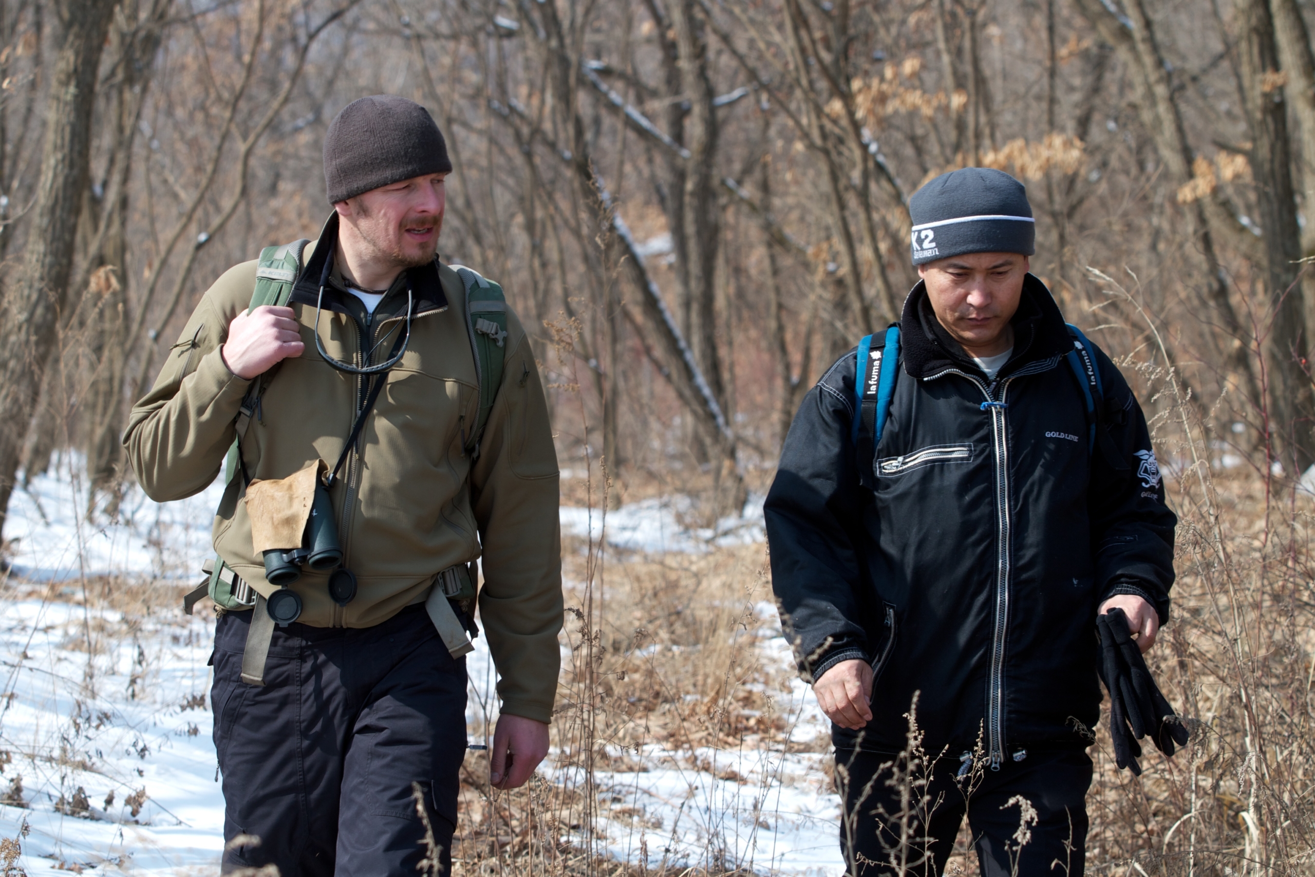 Two men walking through a snowy forest path. One, in a green jacket and hat, peers through binoculars as if searching for a tiger, while the other, clad in black, captures the moment with his camera. Bare trees surround them under a clear sky.