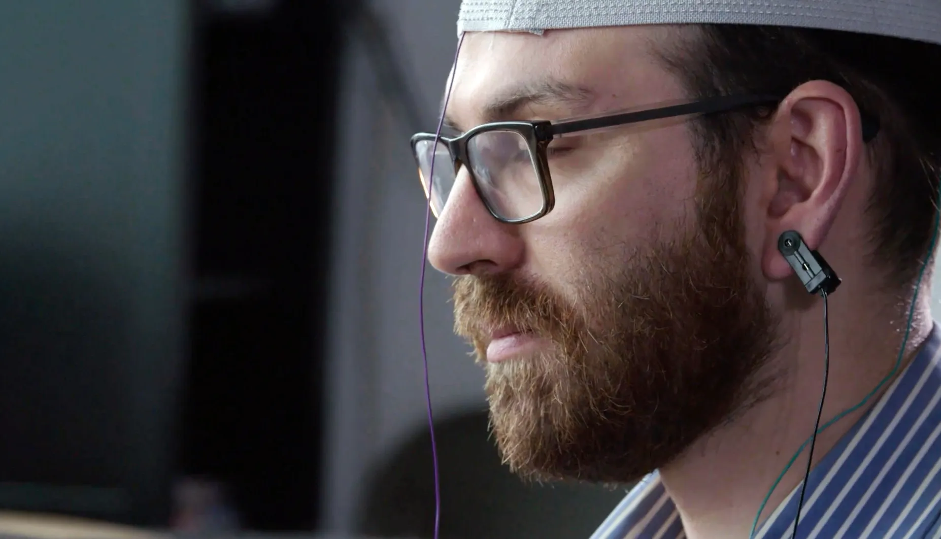 A man with a beard and glasses dons a white cap, likely for the SuperSapiens study, with sensors affixed to his head and ear. He appears focused, possibly engaged in an experiment exploring mind activity.