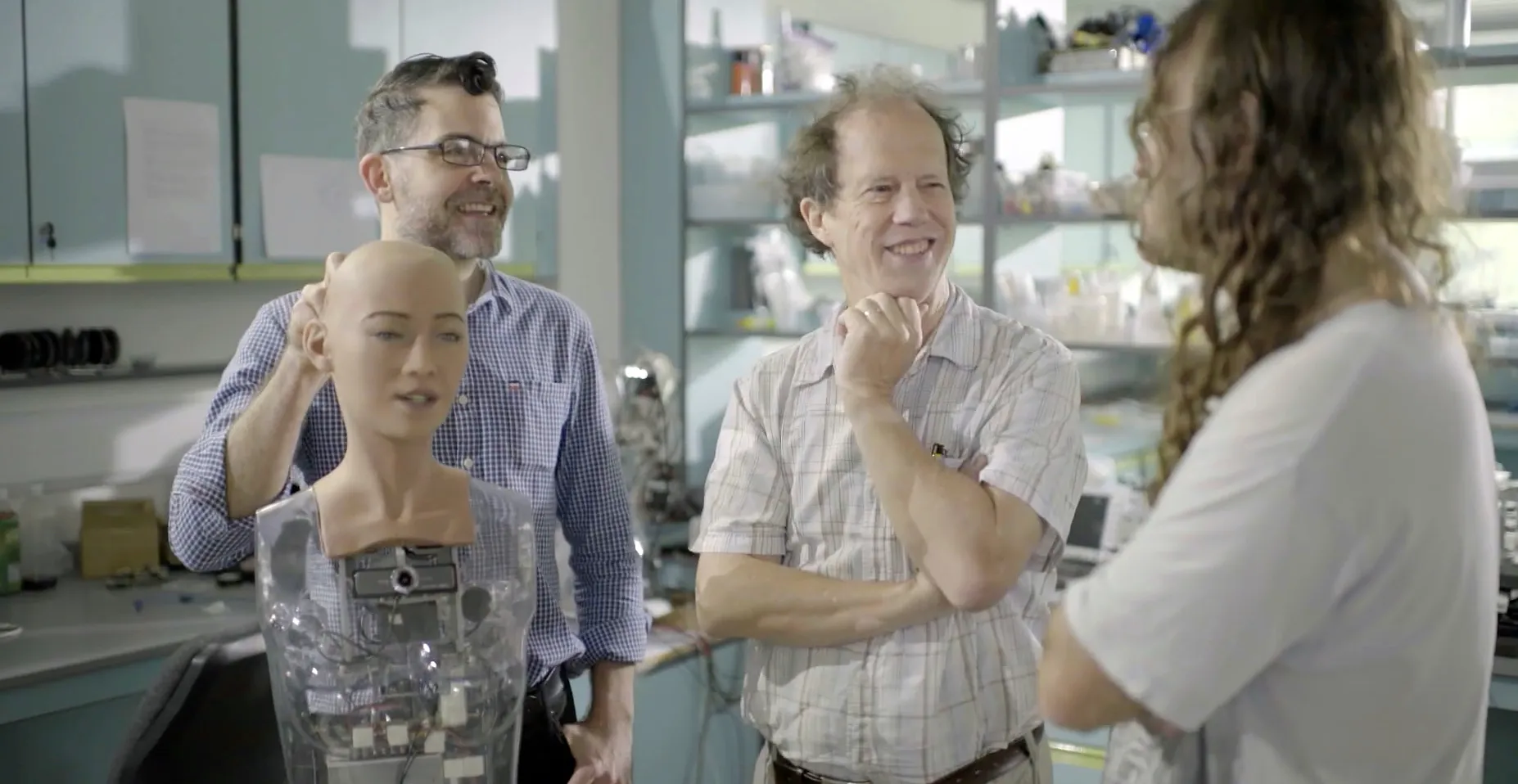 A group of three people stand in a lab with a humanoid robot. The scene captures two men smiling—one with glasses, the other with his hand on his chin. As the third person, their long hair facing away, ponders the rise of SuperSapiens, the robot stands bald and partially transparent.