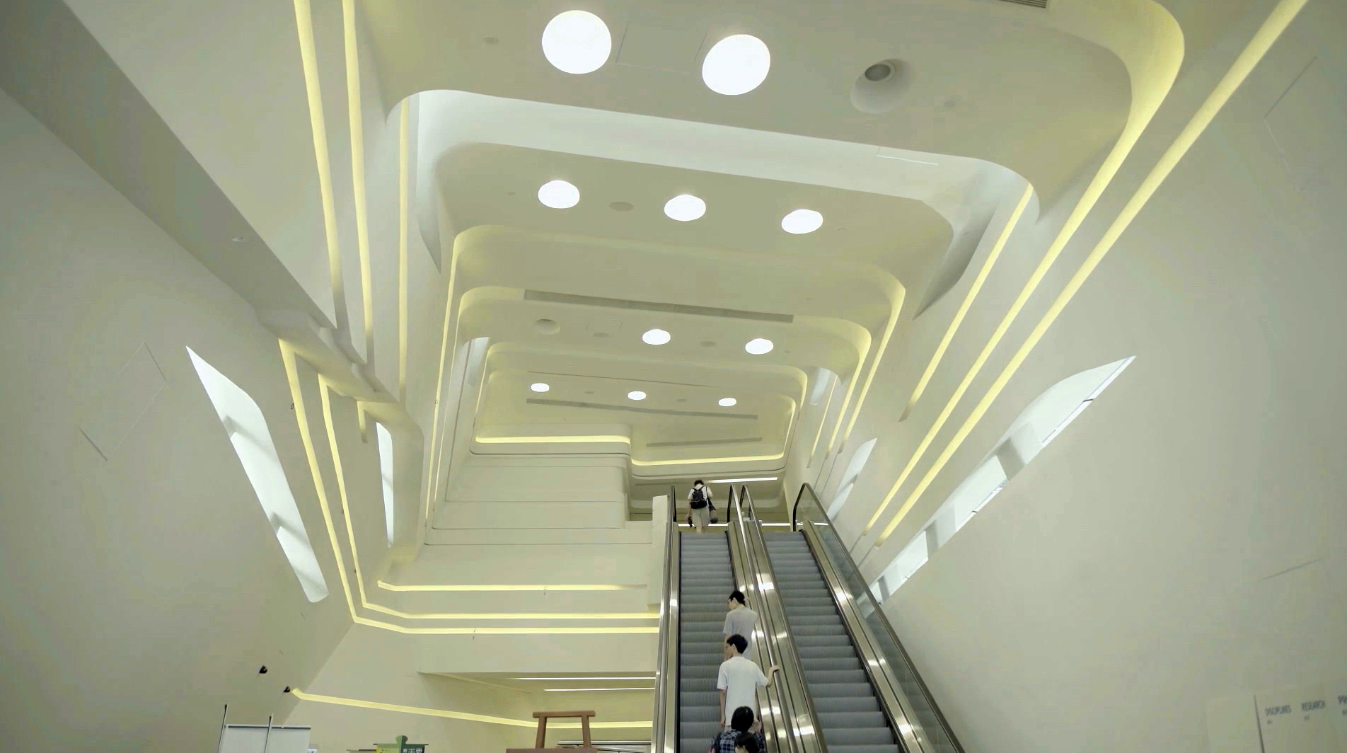 Futuristic, white interior with soft lighting and circular skylights. As the escalator ascends with a few people, echoing the 