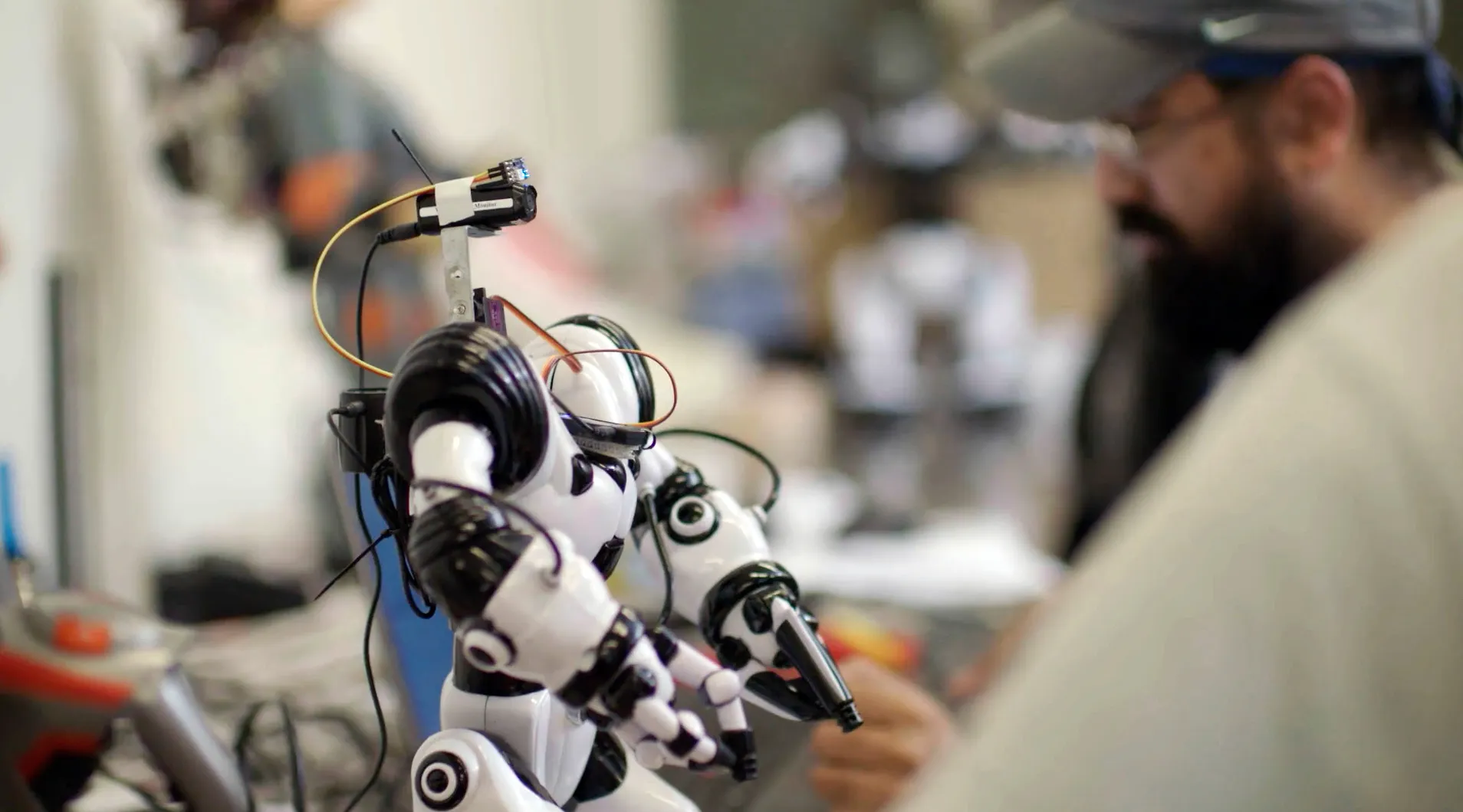 In a workshop setting, a person meticulously works on a small humanoid robot. The focus is on the black and white robot with visible wires and mechanical joints, embodying the rise of SuperSapiens while the blurred figure in the background tinkers with innovation.