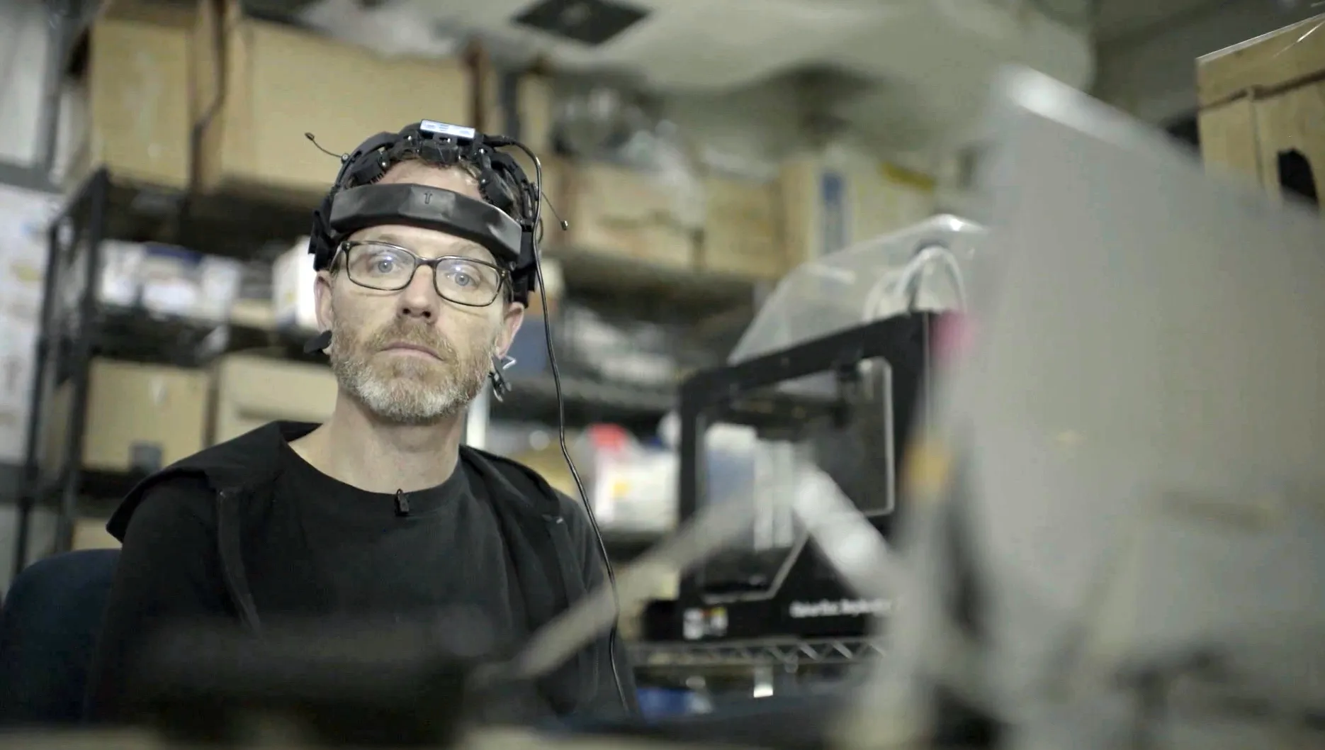 In a cluttered workshop resembling a tech lab, a man wearing glasses and a head-mounted device labeled 