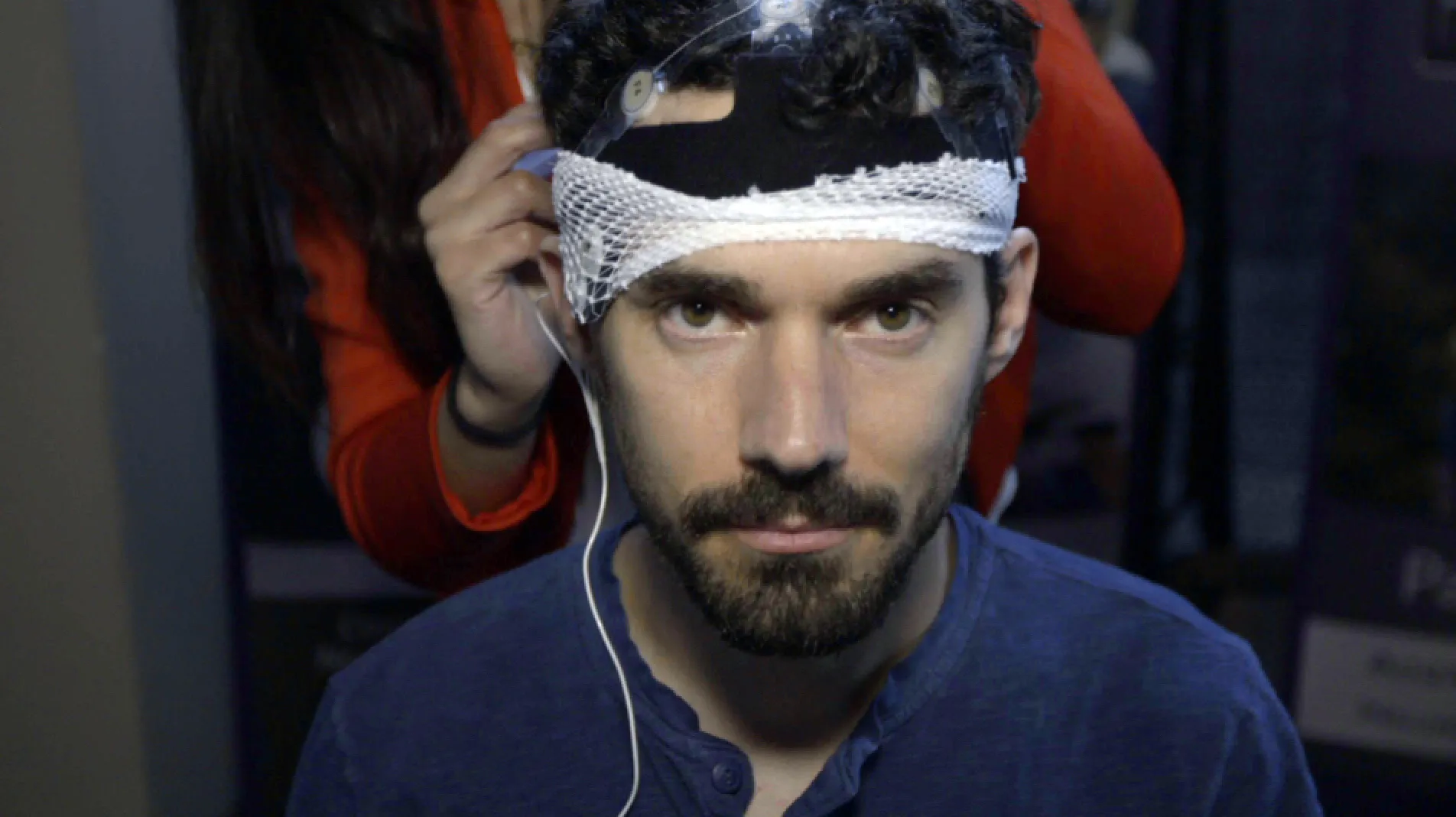 A man with curly hair and a beard wears a headband equipped with SuperSapiens sensors attached to his forehead. A person in a red sleeve carefully adjusts the equipment. The dimly lit background adds an air of intrigue to this exploration of the mind's rise.