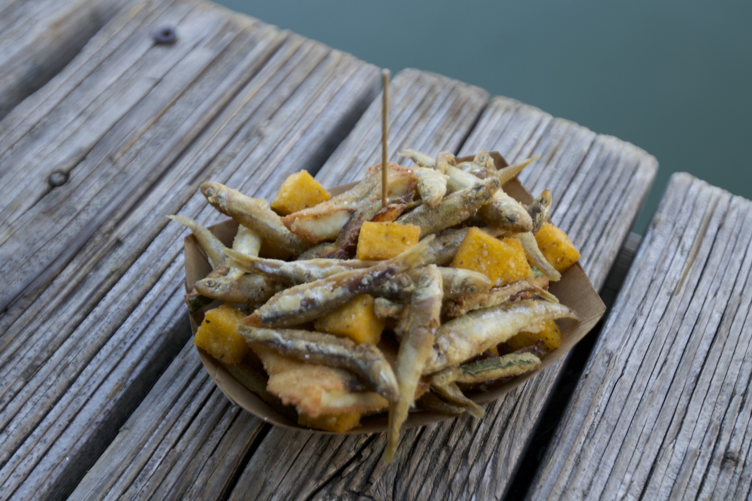 A paper bowl brimming with crispy fried small fish and golden-brown polenta cubes rests on a rustic wooden surface, its vibrant textures popping against the blue-green backdrop.