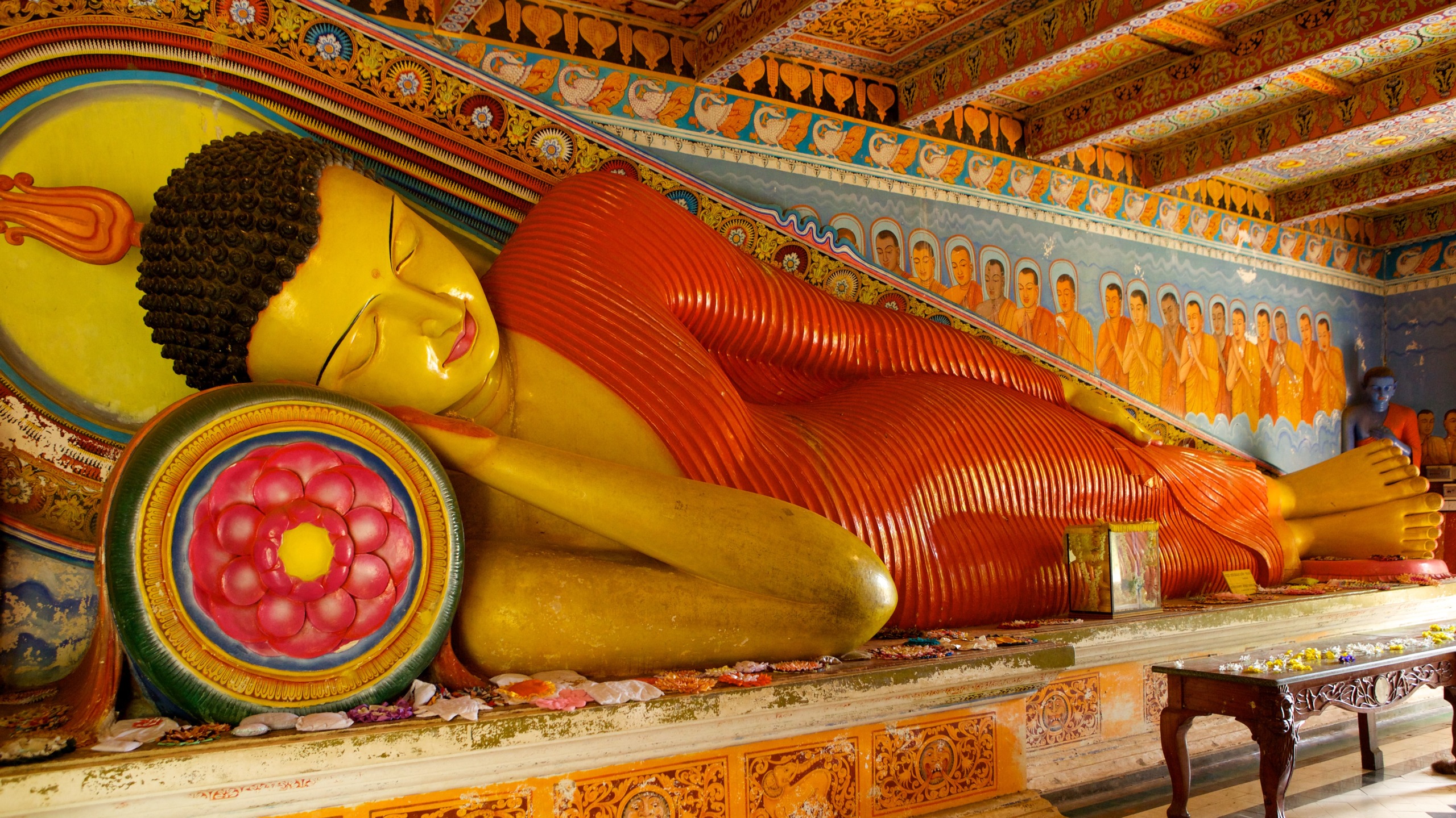 A vibrant statue of a reclining Buddha, adorned in a red robe with a serene expression, rests on a lotus-patterned pillow. Set in an ornately decorated Sri Lankan temple, the background features intricate murals and an array of figures.