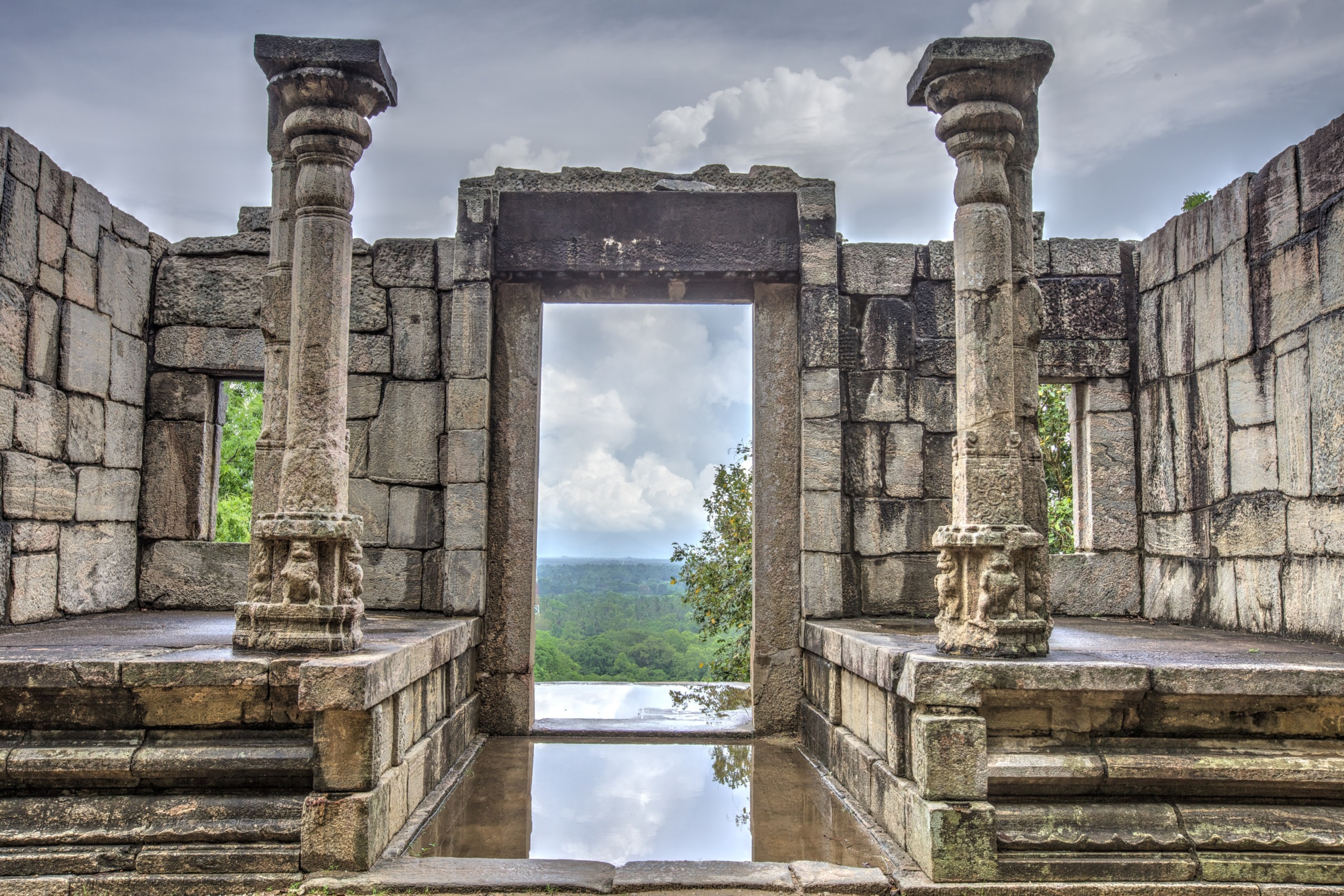 An ancient stone structure with two ornate pillars and a central doorway offers a view reminiscent of Sri Lanka's lush green forests and cloudy skies. The wet floor mirrors parts of the architecture and the sky above, evoking a sense of serene beauty.