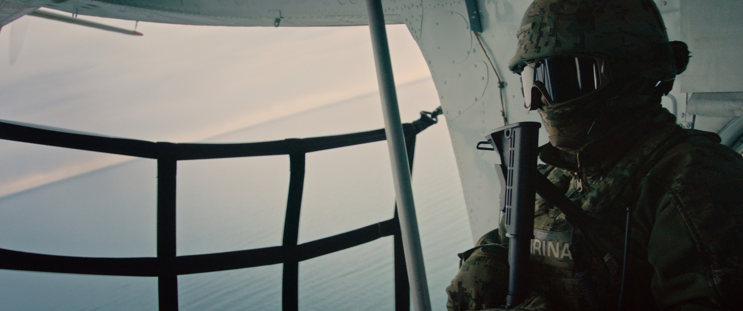 A soldier in camouflage and tactical goggles sits at the open door of a military aircraft, gazing over a sea of shadows. The aircraft's structure looms behind him, the scene bathed in muted, cool tones.