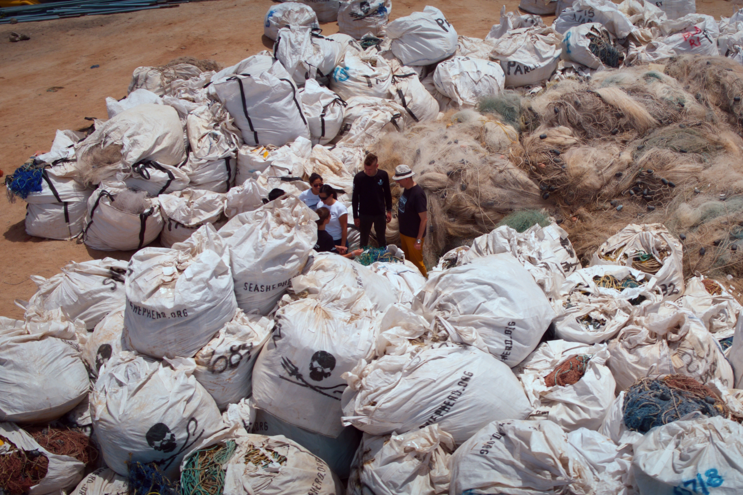 From an aerial viewpoint, a sea of shadows unfolds as large piles of discarded fishing nets, encased in white bags marked with skull and crossbones symbols, scatter across the sandy coastal ground. Several people navigate through this haunting scene, examining the remnants left behind.