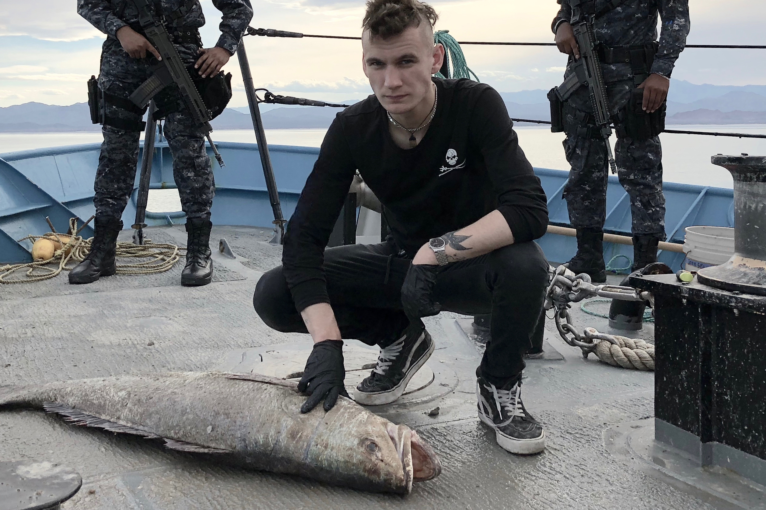 A person in black clothing and gloves crouches on a ship deck beside a large fish, surrounded by the ominous sea of shadows. Armed guards stand in the background as distant mountains loom beyond the vessel.