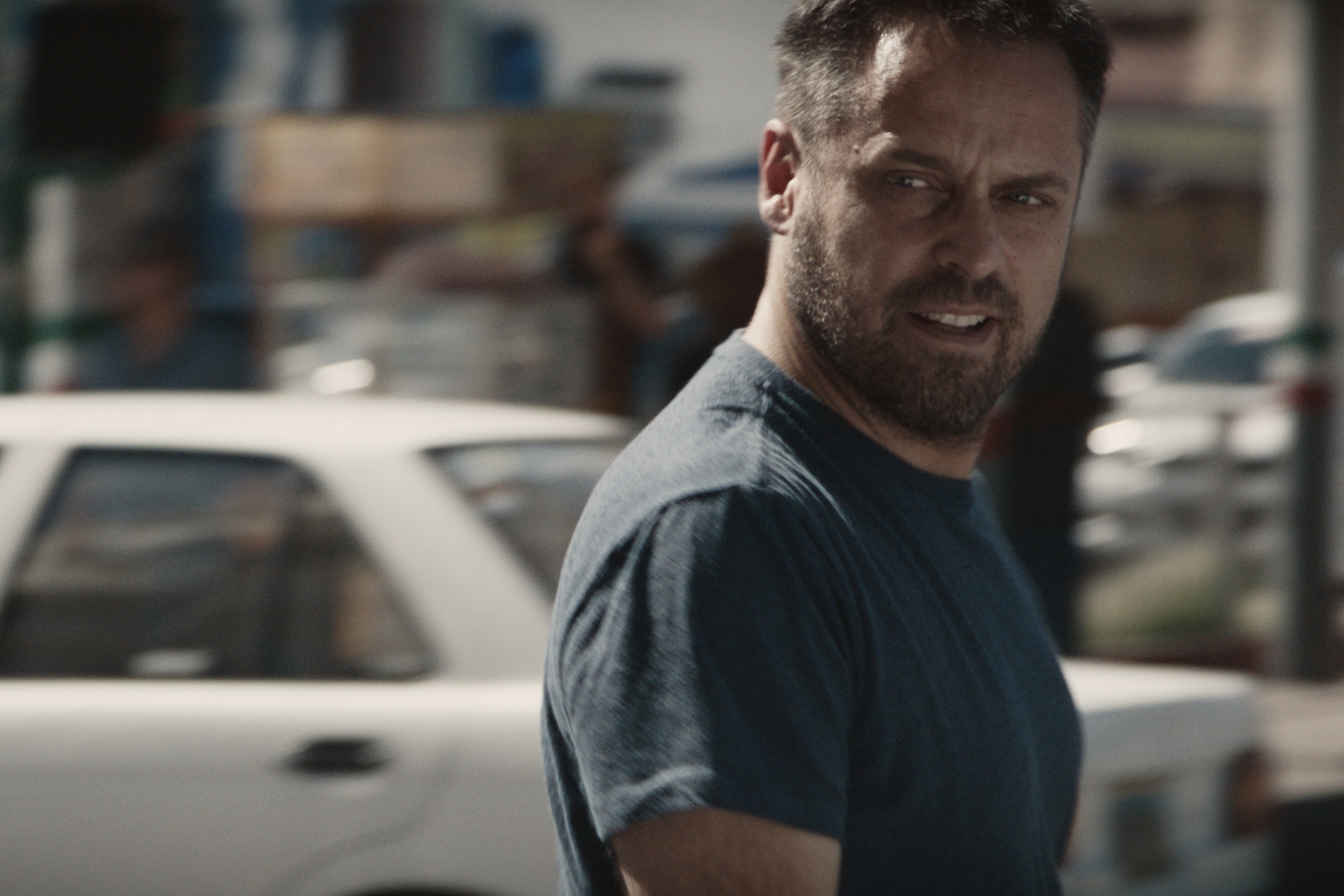 A man with a beard and a blue t-shirt stands outdoors, gazing to the side. Amidst the sunny, bustling scene with blurred buildings and a white car, he seems like a solitary figure in a sea of shadows.