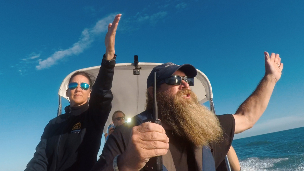 Two people are in a boat on the water. The man with a long beard and sunglasses is steering, arms wide open. The woman beside him raises one arm. Both wear sunglasses and jackets as they navigate the sea of shadows under a clear blue sky, with endless ocean in the background.