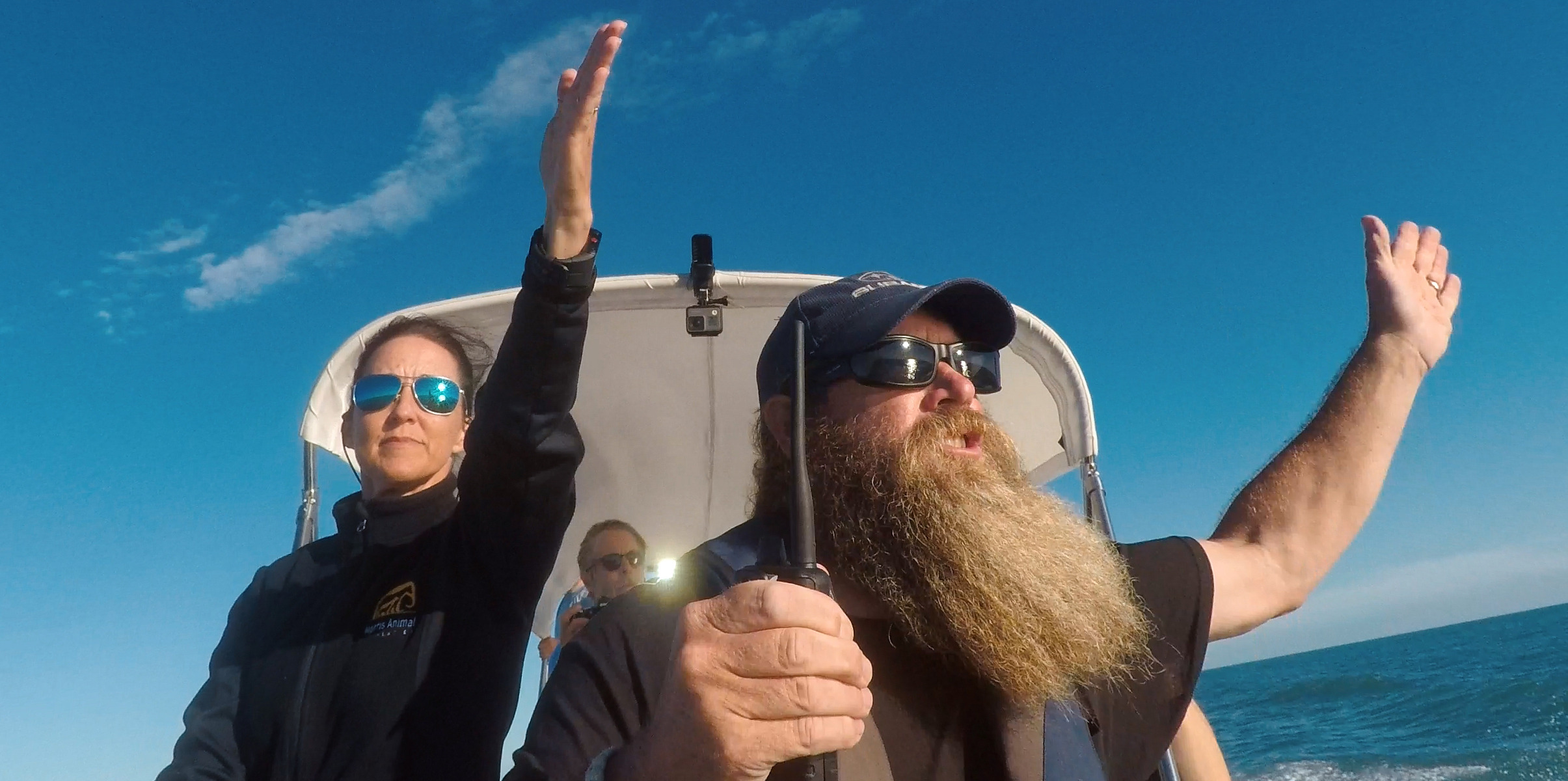 Two people are in a boat on the water. The man with a long beard and sunglasses is steering, arms wide open. The woman beside him raises one arm. Both wear sunglasses and jackets as they navigate the sea of shadows under a clear blue sky, with endless ocean in the background.