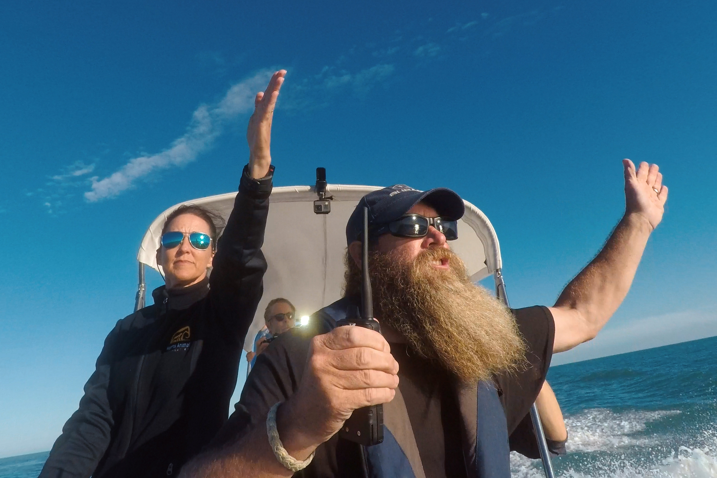 Two people are in a boat on the water. The man with a long beard and sunglasses is steering, arms wide open. The woman beside him raises one arm. Both wear sunglasses and jackets as they navigate the sea of shadows under a clear blue sky, with endless ocean in the background.