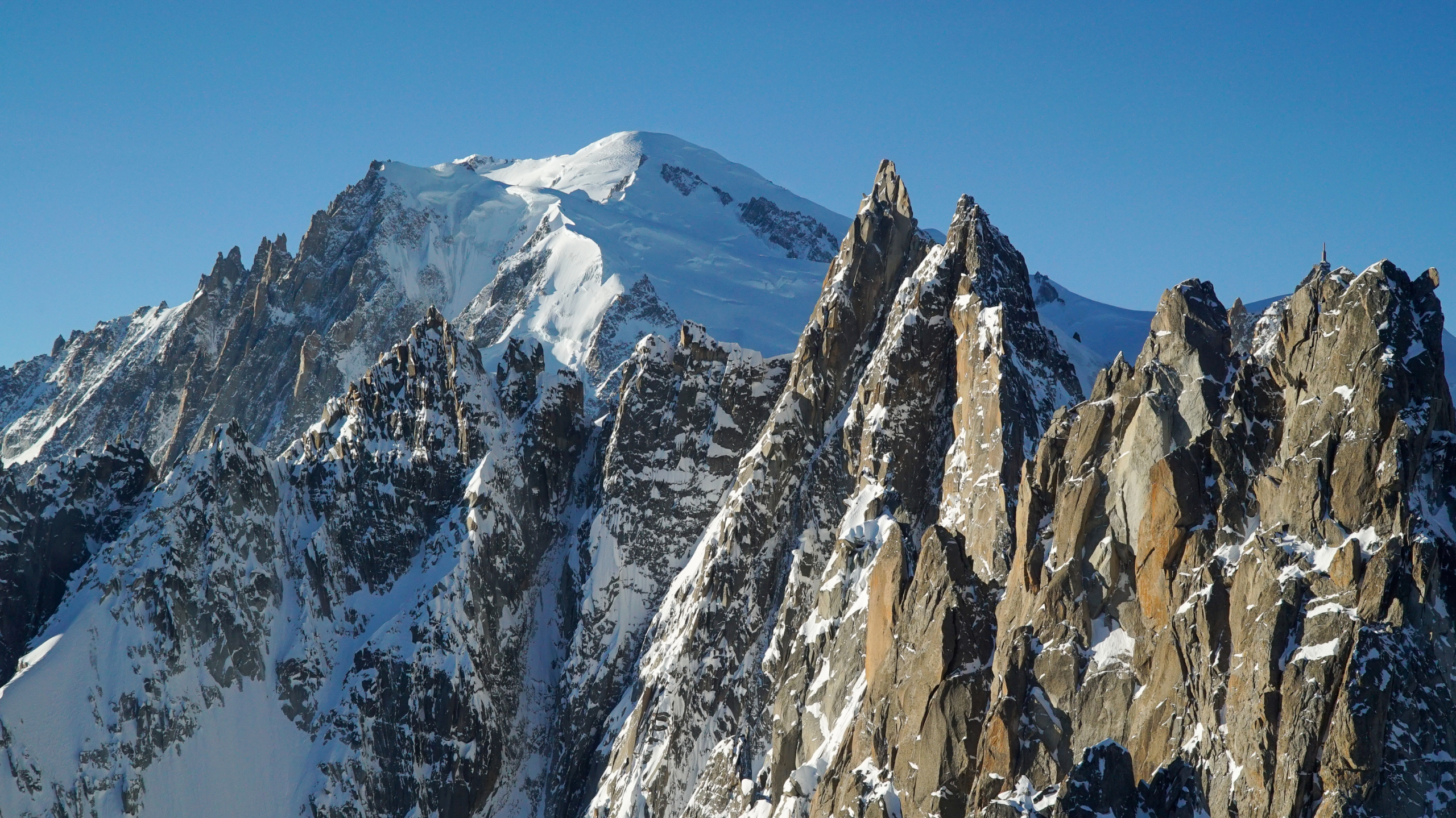 Snow-covered mountain peaks with jagged rocks are bathed in sunlight under a clear blue sky, showcasing a dramatic and rugged alpine landscape reminiscent of the majestic Alps.