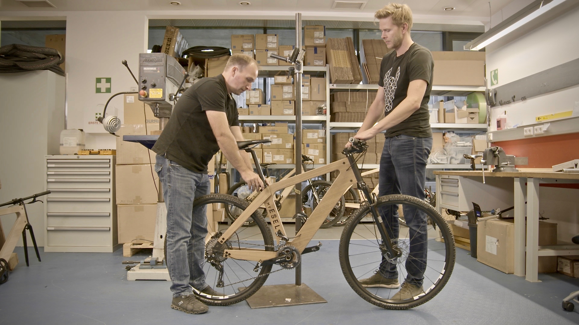 In a workshop rooted in craftsmanship, two men examine a wooden bicycle frame. Surrounded by shelves filled with boxes and equipment, they focus on adjusting the bike. The space tells a story of creativity—cluttered but organized like the branches of towering trees.