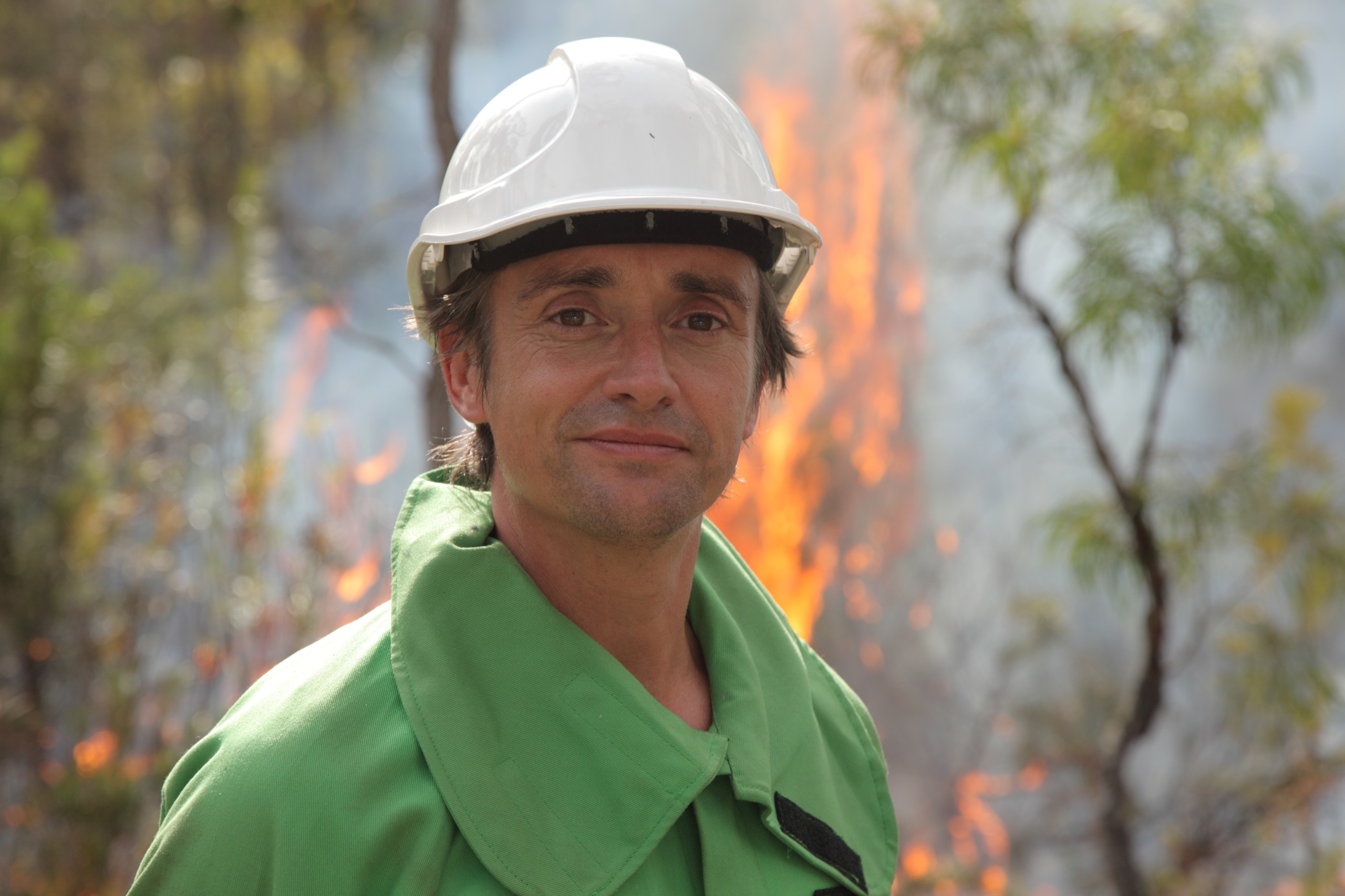 A person in a green jacket and white hard hat stands in front of a small, controlled fire, reminiscent of a scene from Wild Weather with Richard Hammond, set against a backdrop of trees and drifting smoke in the forested area.