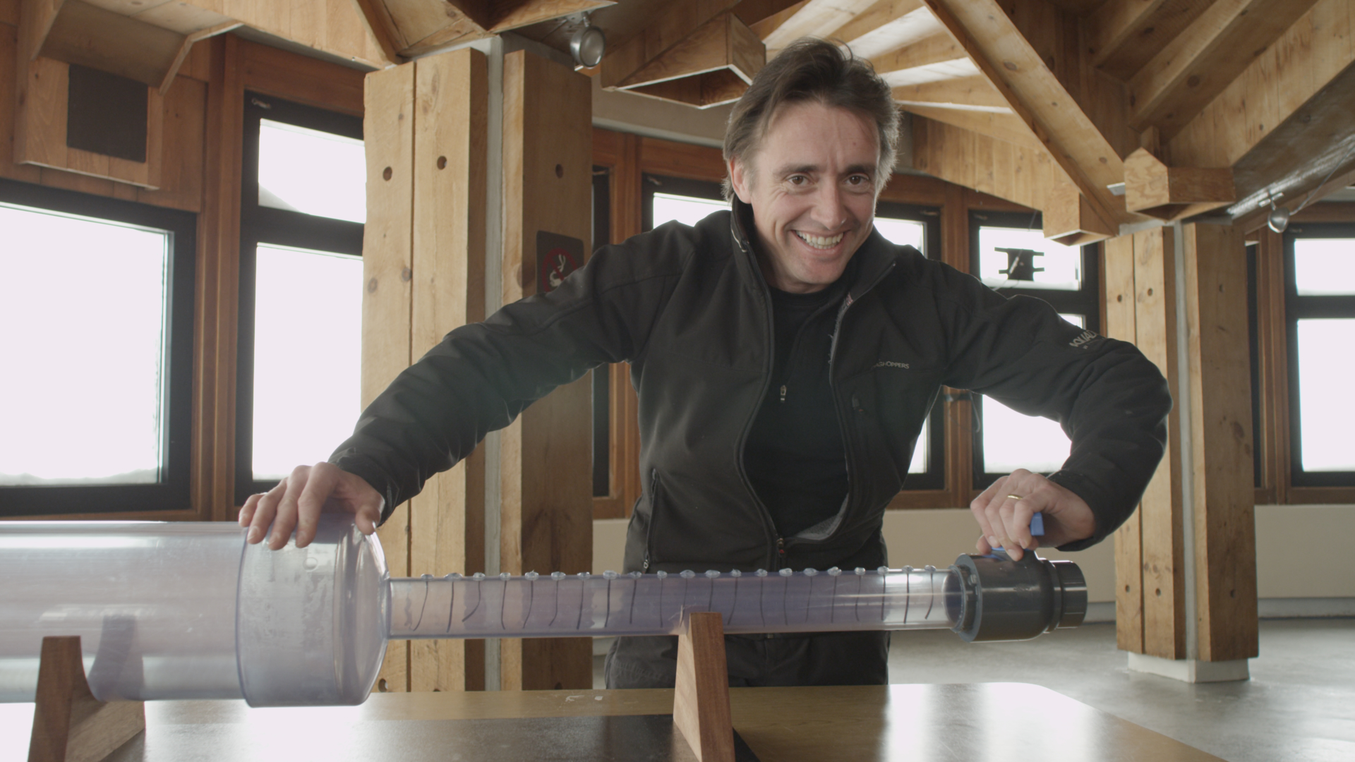 A person smiles while engaging with a long, transparent plastic tube placed on wooden supports in a room with large windows and wooden beams, reminiscent of Richard Hammond's Wild Weather explorations.