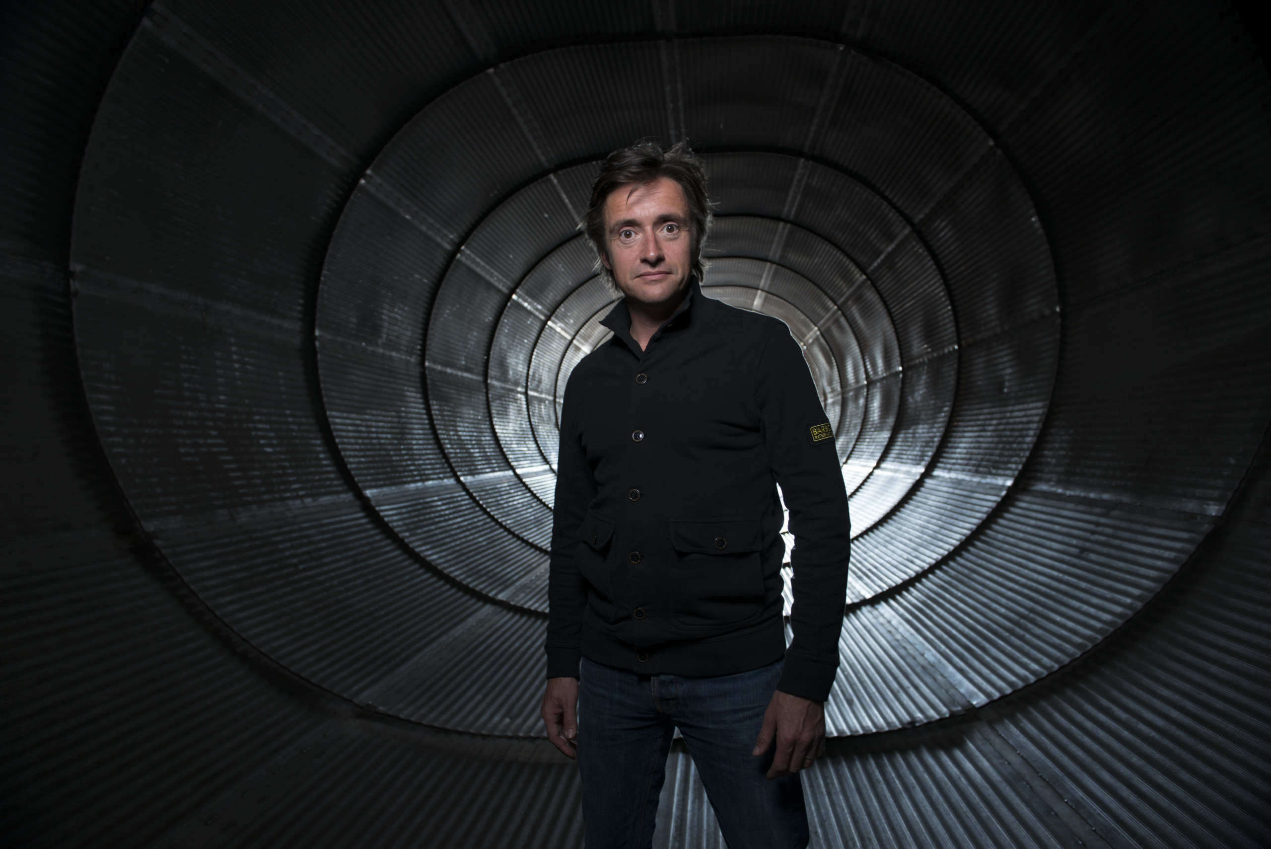 A person stands in a metallic tunnel, wearing a dark jacket and jeans. The tunnel's circular pattern creates depth, and soft lighting highlights their features—an atmosphere that could be straight out of a Wild Weather documentary with Richard Hammond.