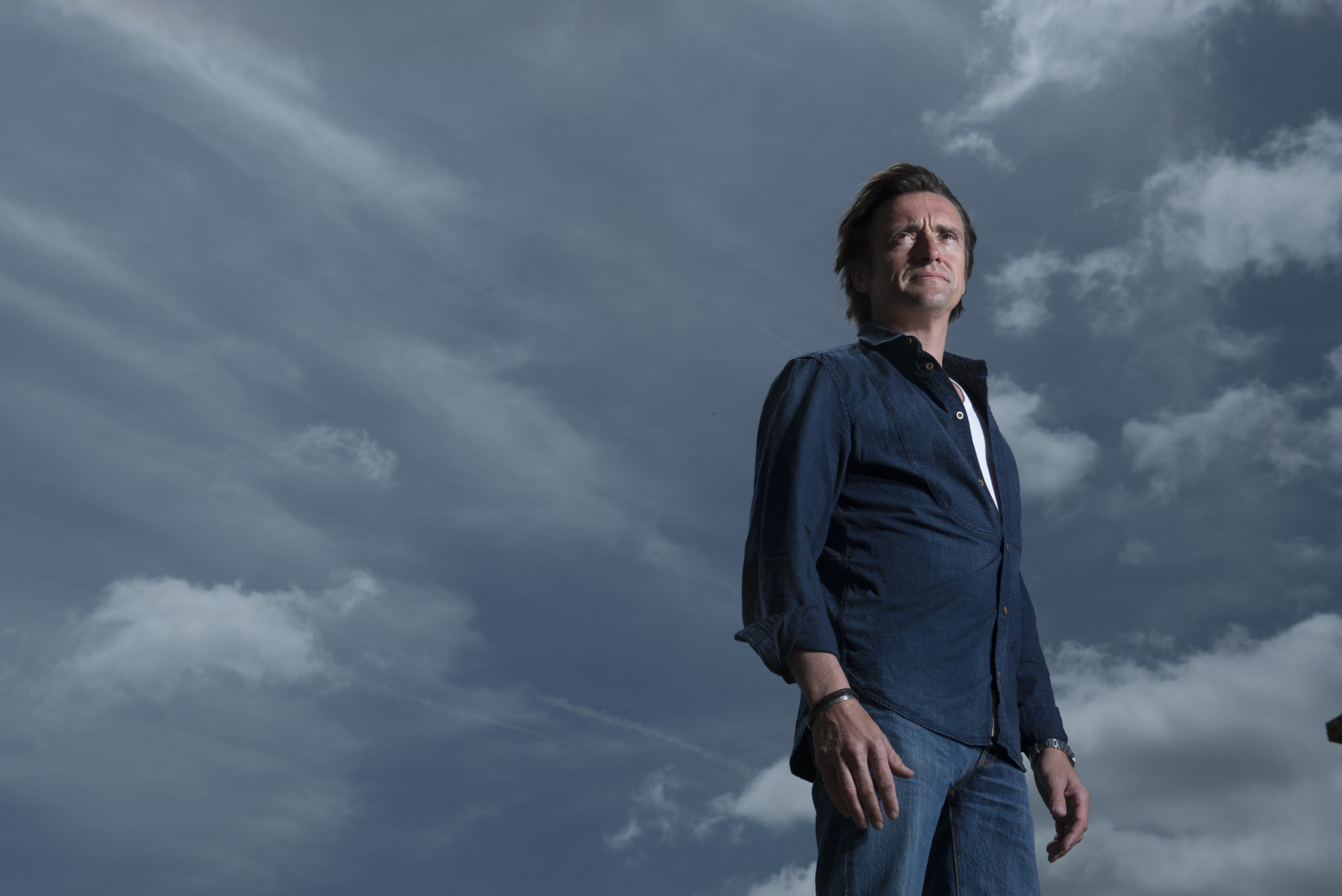 A person in a blue shirt and jeans stands outdoors under a cloudy sky, reminiscent of Richard Hammond exploring wild weather, gazing into the distance with their hands by their sides.