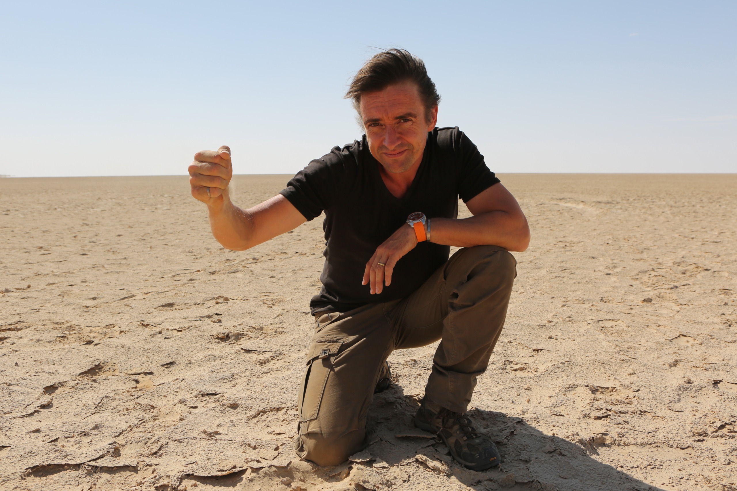 A person wearing a black shirt and khaki pants kneels on dry, cracked earth in a barren desert landscape, reminiscent of a Wild Weather episode hosted by Richard Hammond. They have a watch on their left wrist and are gesturing with their right hand under the clear blue sky.