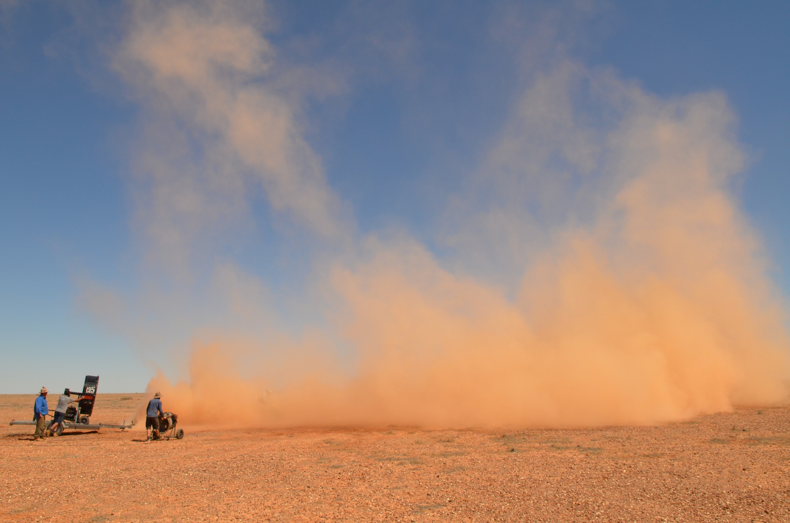 A vast desert landscape under a clear blue sky hosts a wild weather spectacle as a massive dust cloud sweeps across the ground. To the left, two people with equipment stand on the dry, reddish-brown earth, capturing footage for a gripping weather documentary.