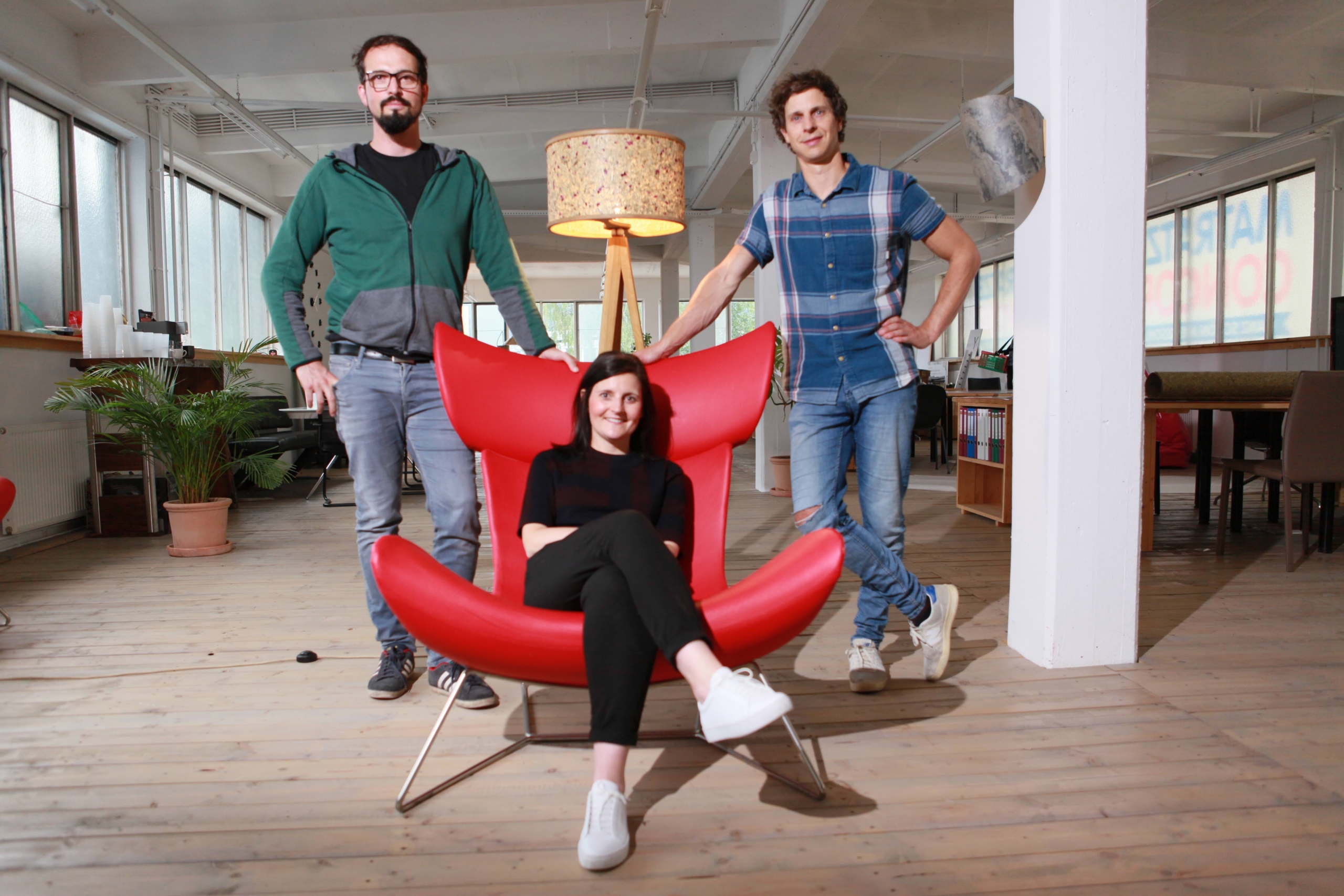 Three people in a modern office: a woman sits in a red chair, smiling, while two men stand behind her. The space features wooden floors, large windows, a tall lamp, and plants.