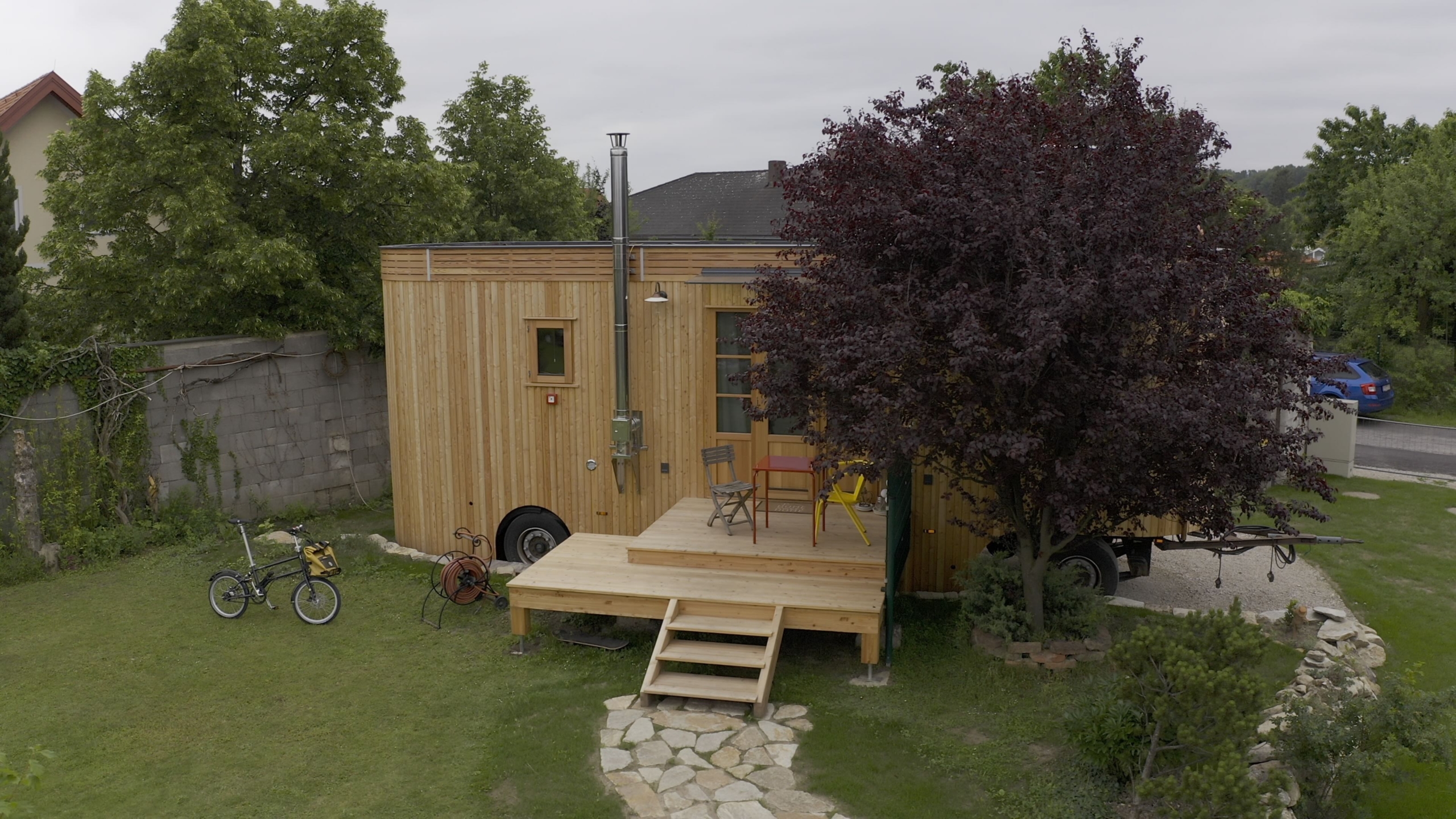 Wooden tiny house on wheels with a small porch, surrounded by trees and grass. A bicycle and a wheelbarrow are on the lawn. A stone path leads to the steps.