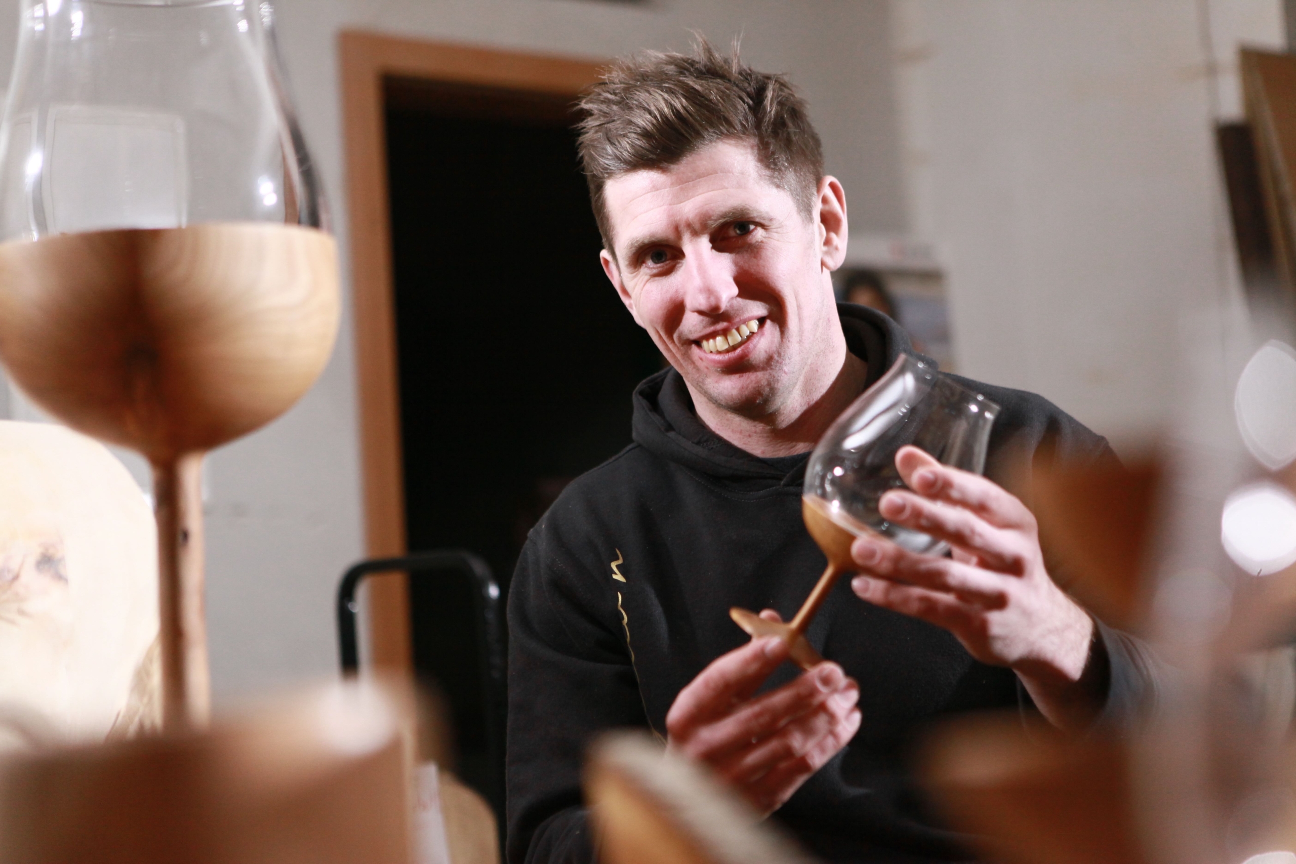 A person smiles while holding a wooden object, surrounded by similar items on a table. They are indoors, wearing a black hoodie, with a wooden door and wall in the background.