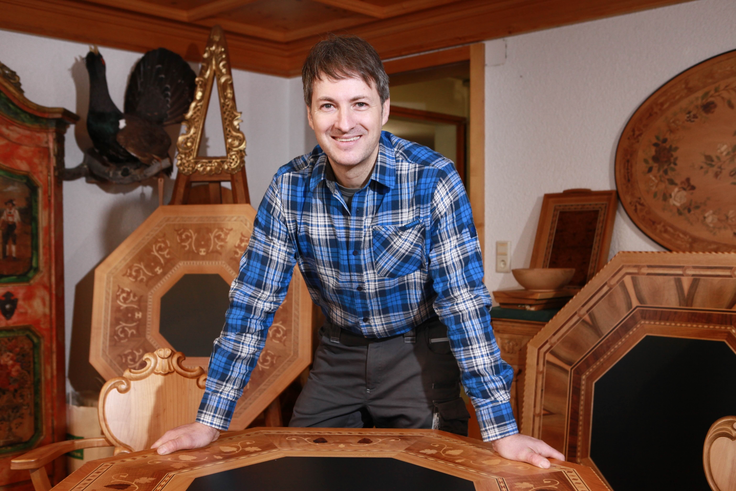 A person in a blue plaid shirt is smiling, leaning forward with hands on a wooden table. Behind them are intricately designed wooden frames and items, suggesting a woodworking or artisan setting.