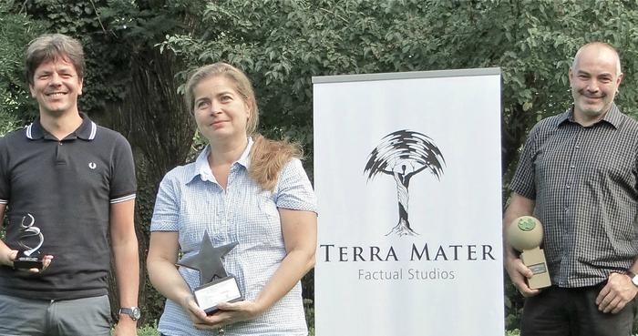 Three people stand outdoors, smiling and holding trophies, as a banner for Terra Mater Factual Studios hangs behind them. Trees and greenery surround the winners, whose joy reminds us of the infectious energy found in hits of 2012.