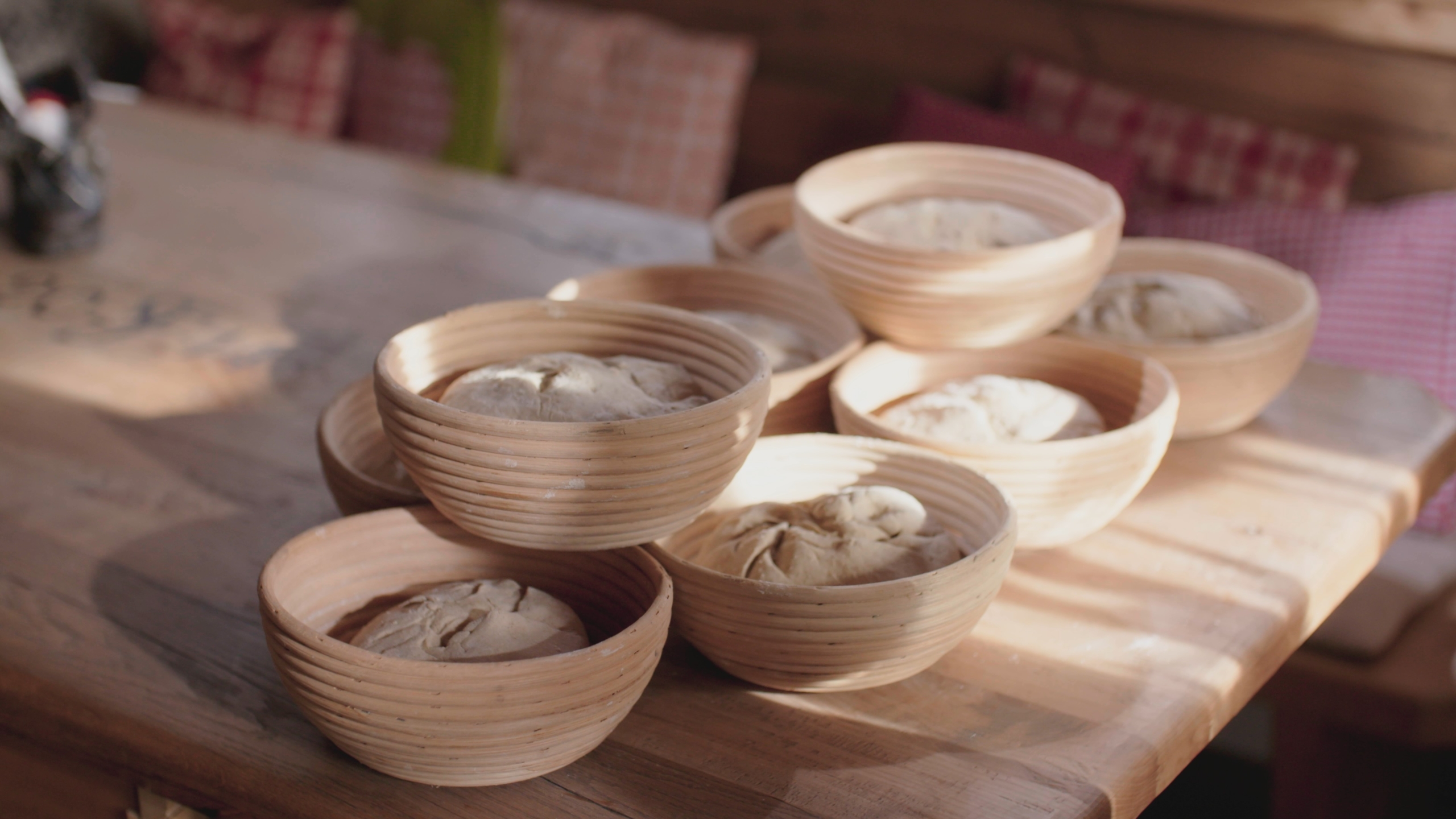 Wooden bowls filled with rising dough are arranged on a sunlit wooden table, capturing the essence of the lights of home. The scene suggests a warm, rustic kitchen setting, with soft pillows in the background adding to the cozy ambiance.