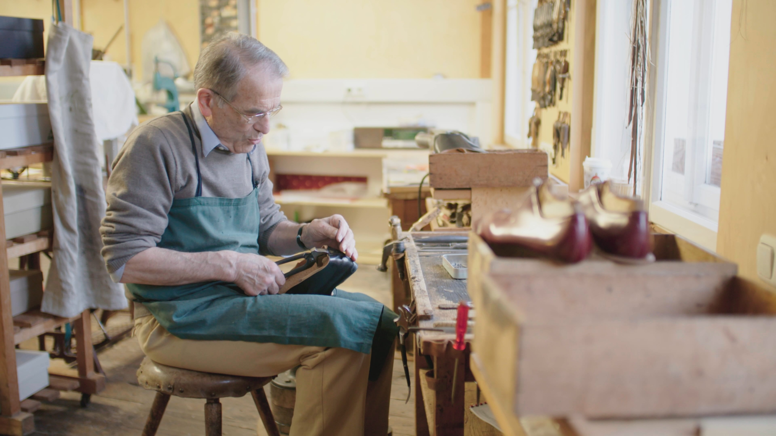 An elderly man in an apron works diligently on a shoe at a cobbler's bench, bathed in the warm glow of lights of home. Tools and shoe lasts are scattered around the wooden workspace, creating an atmosphere of dedication and craftsmanship.