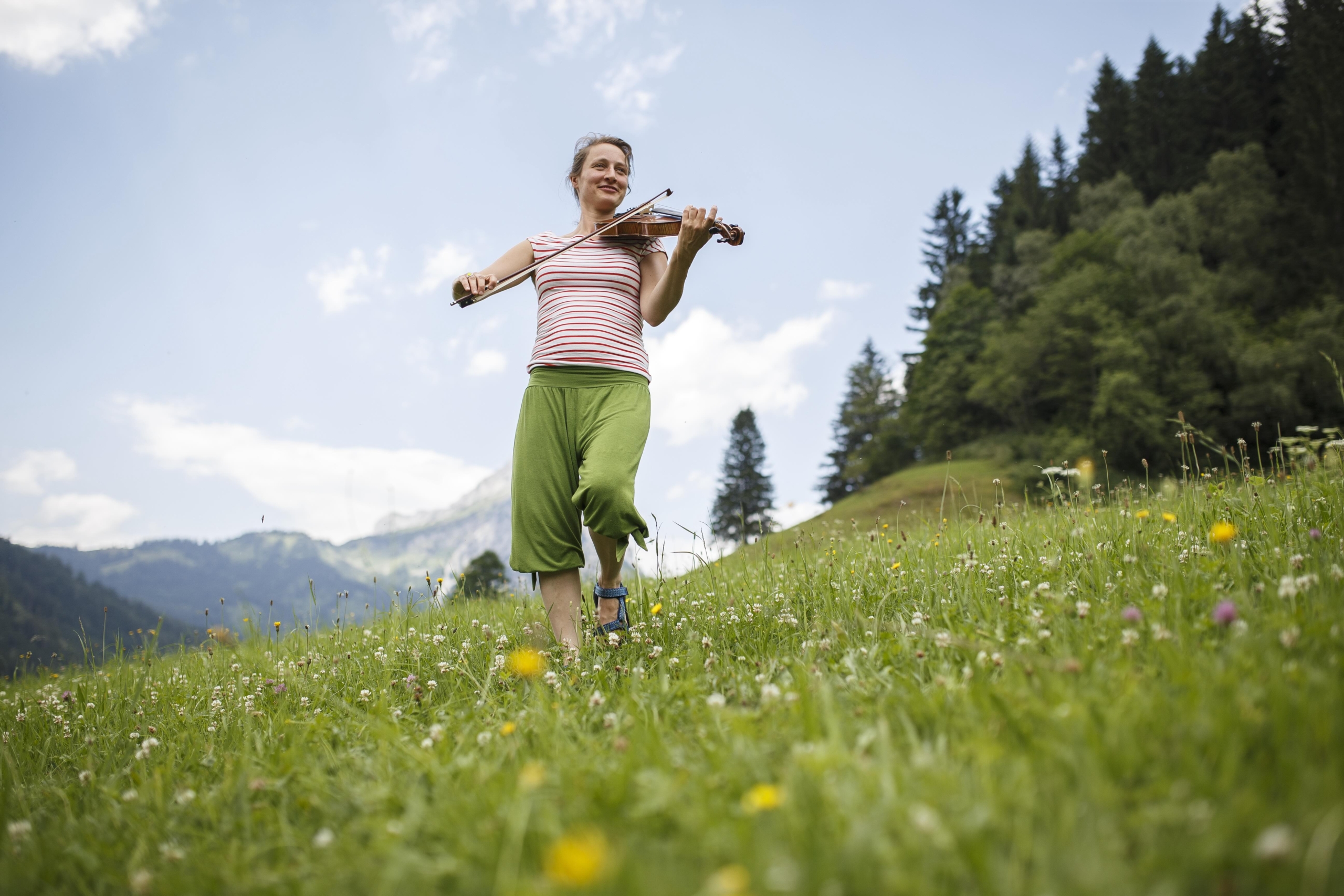 A woman plays the violin, harmonizing with the lights of home as she strolls through a vibrant, flower-filled meadow framed by mountains and pine trees. Dressed in a striped shirt and green pants, she smiles under the clear blue sky.