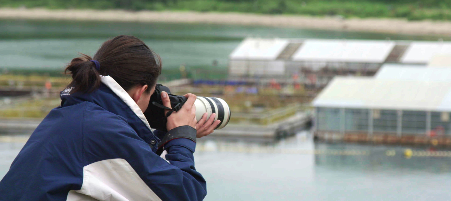 A person with long hair in a ponytail is taking a photo with an orca-sized camera lens. They're wearing a blue and white jacket, standing near a body of water with buildings and trees visible in the background.