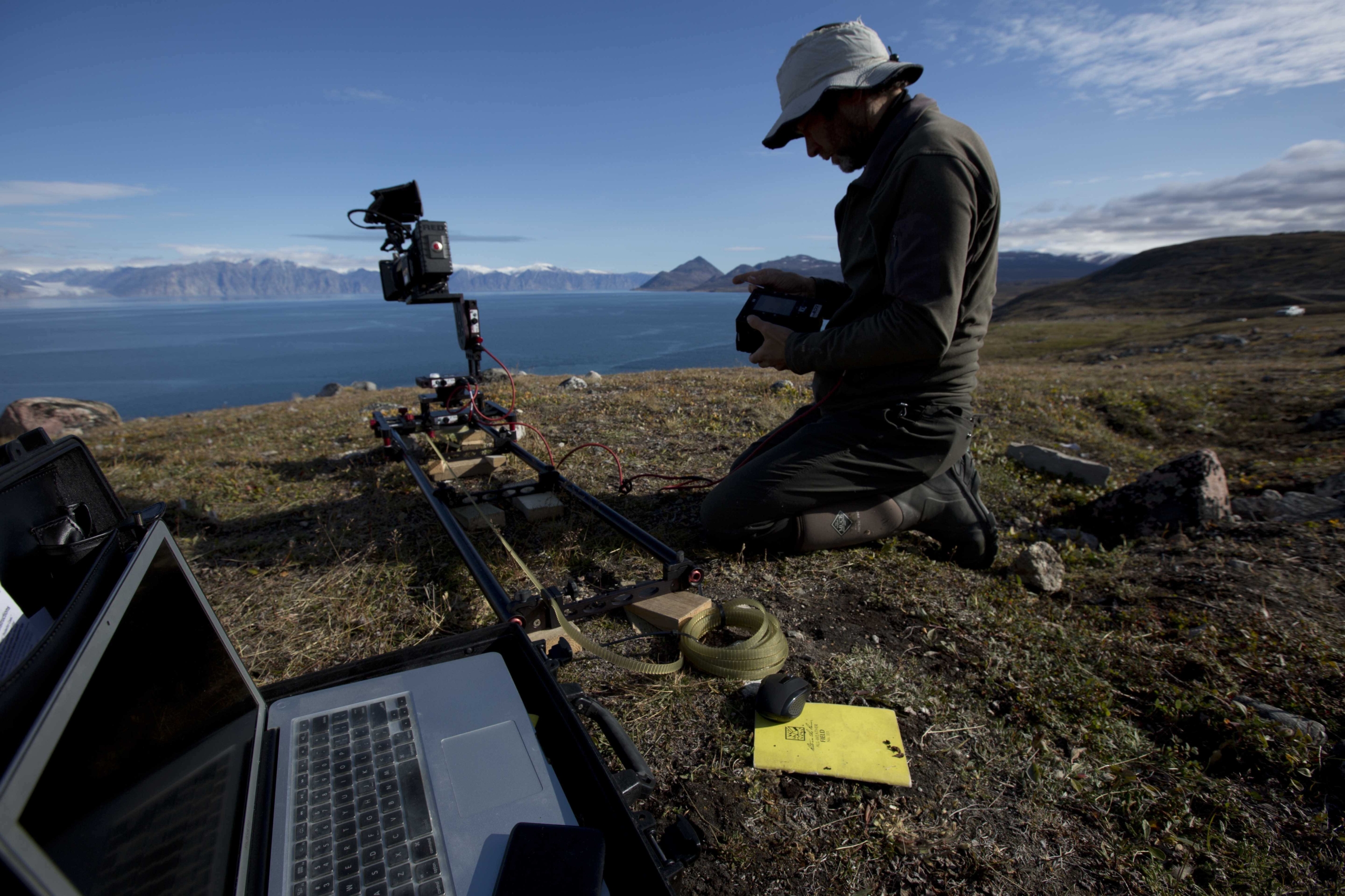 A person in outdoor clothing kneels on grassy terrain by the sea, operating camera equipment on a track. A laptop displaying Terra Mater insights is open nearby. The scene is set against a backdrop of mountains and a partly cloudy sky.